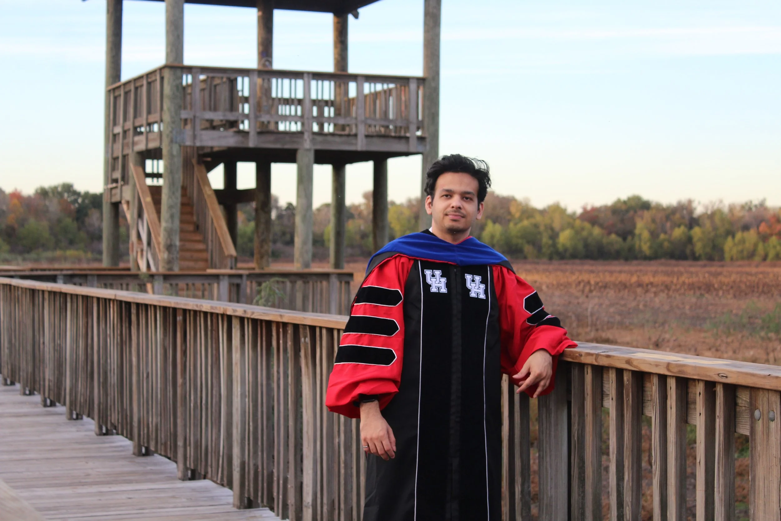 Shahmeer wears PhD graduation robes while standing on a bridge overlooking a field.