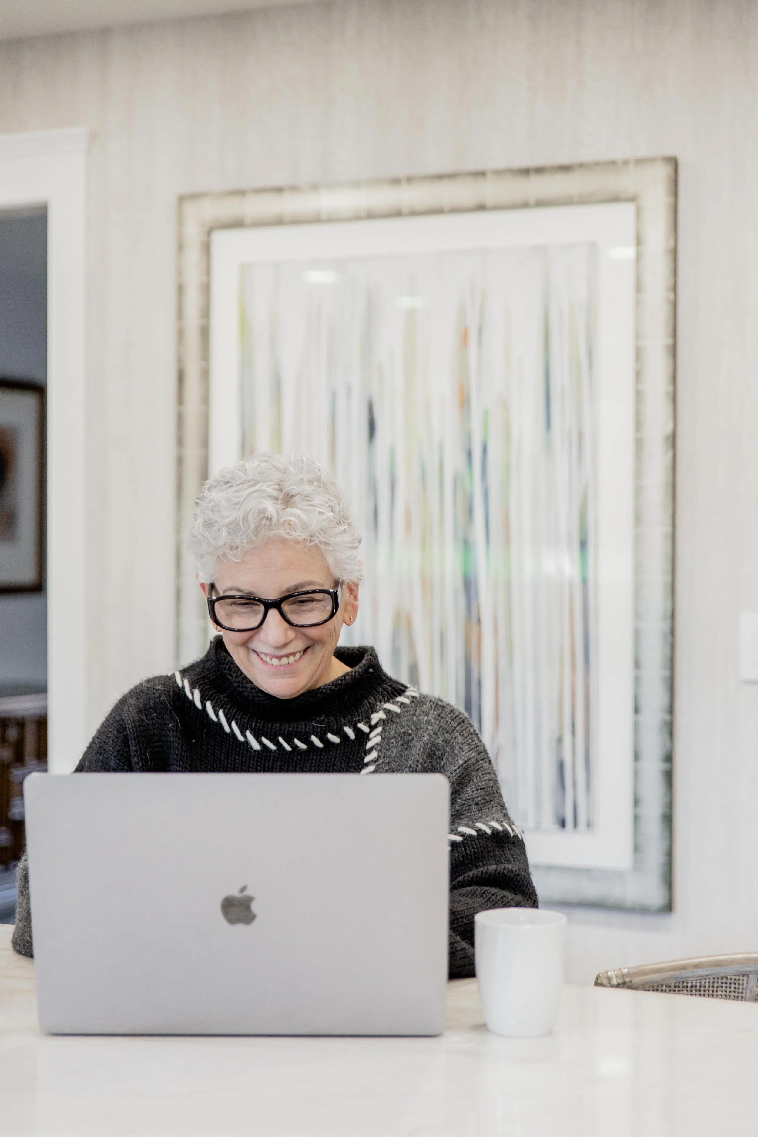 Smiling elderly woman with curly gray hair and glasses using a silver MacBook at a dining table, with a white mug nearby, in a room with a colorful abstract painting in the background.
