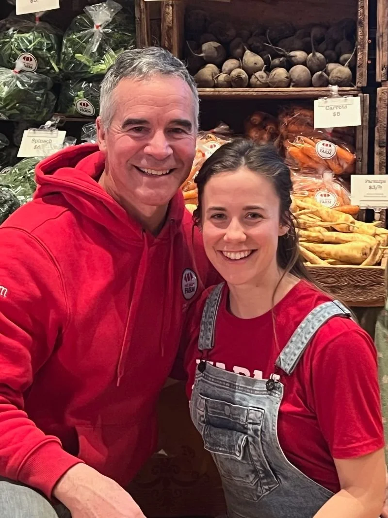 A man in a red hoodie and a woman in overalls and a red shirt smile in front of a produce display at the farm store