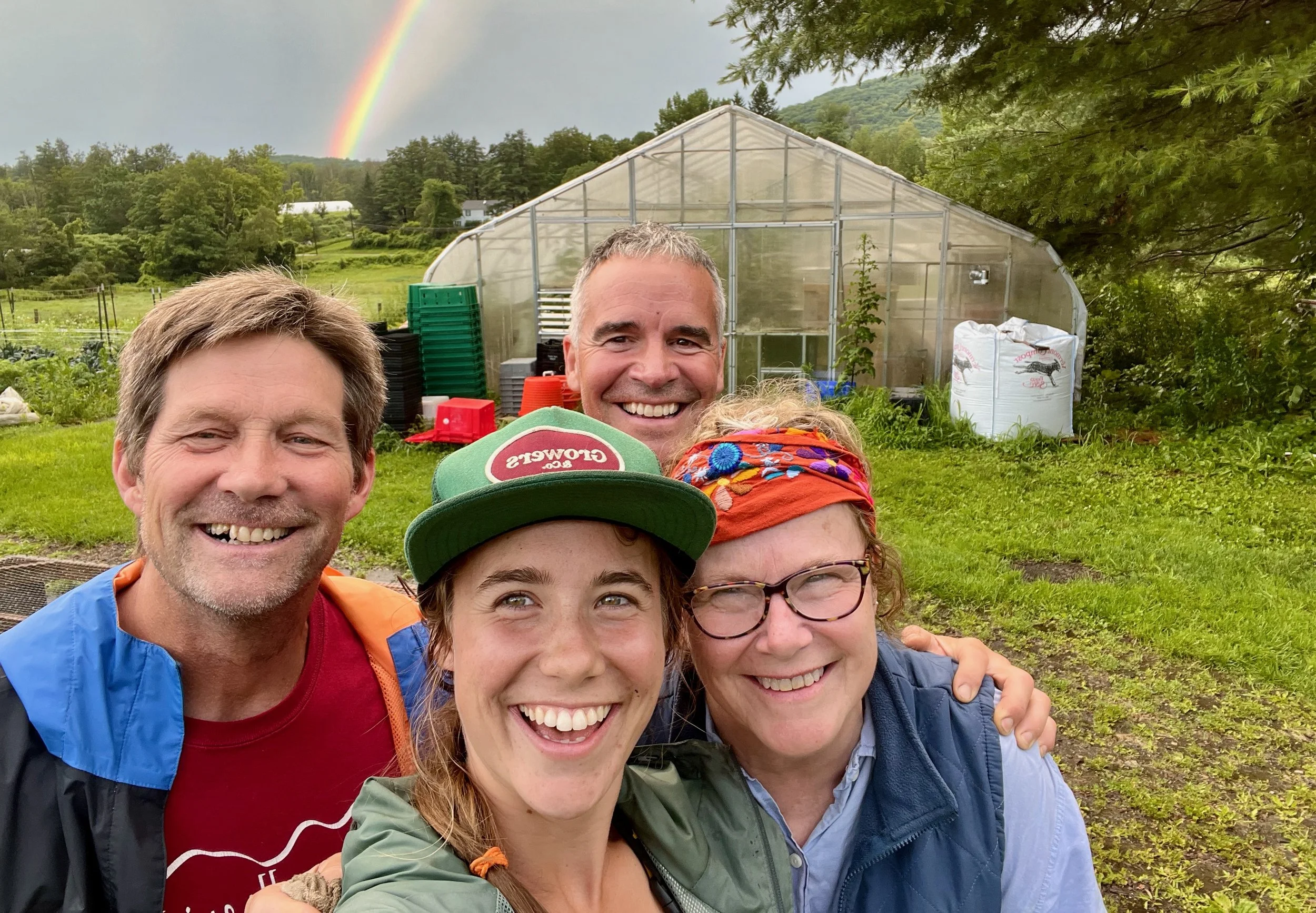 Four people standing close together outdoors, with a greenhouse and a rainbow behind them