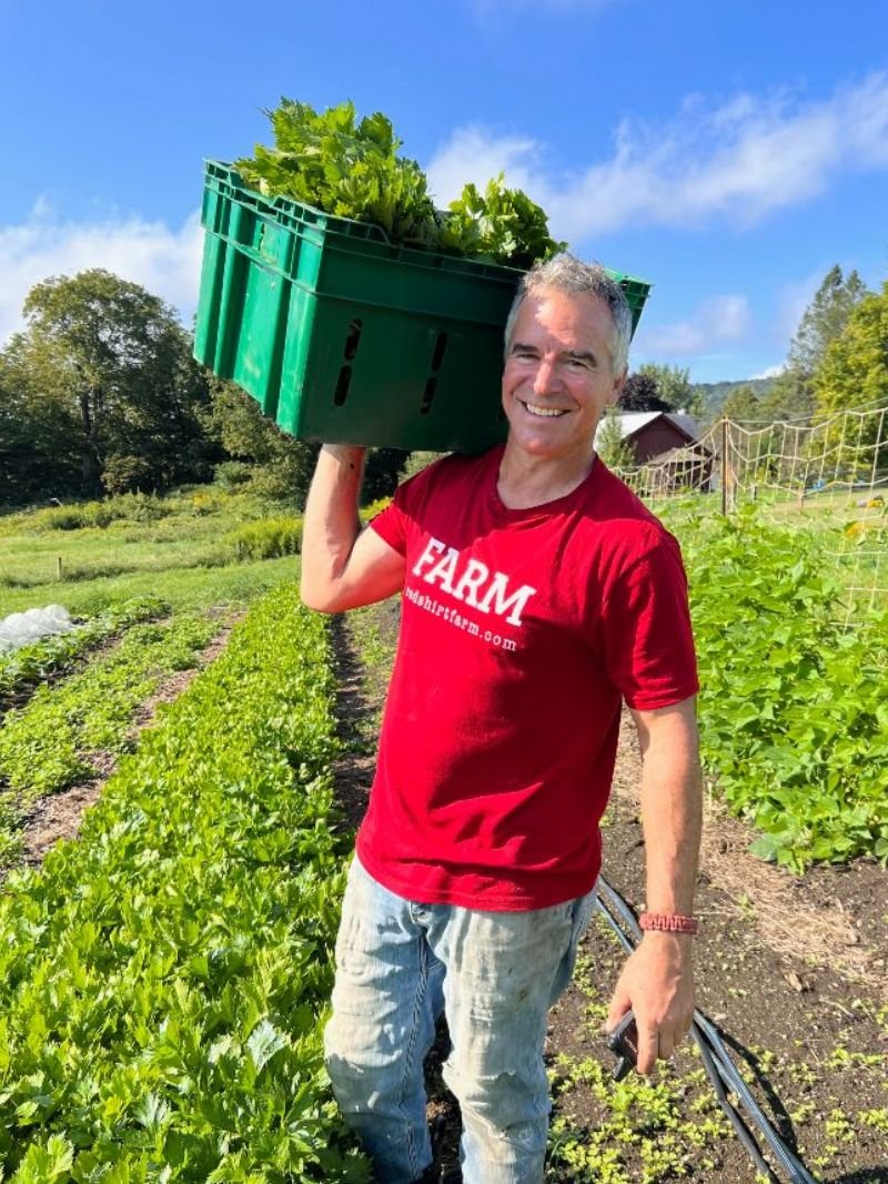 A man in a red FARM t-shirt carries a green crate on his shoulder between rows of vegetables
