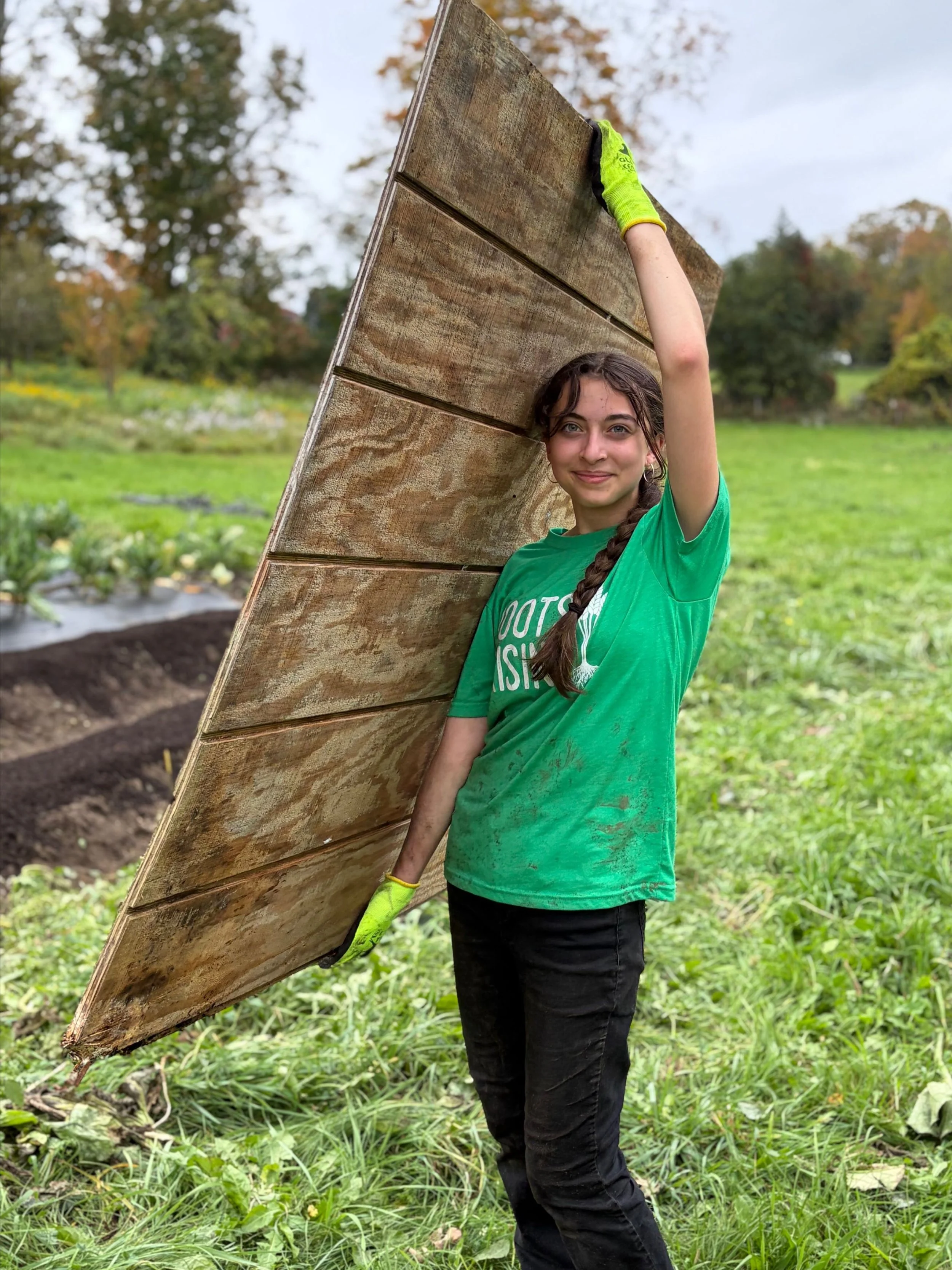 A Roots Rising student carries a wooden board on her shoulder across a harvested field