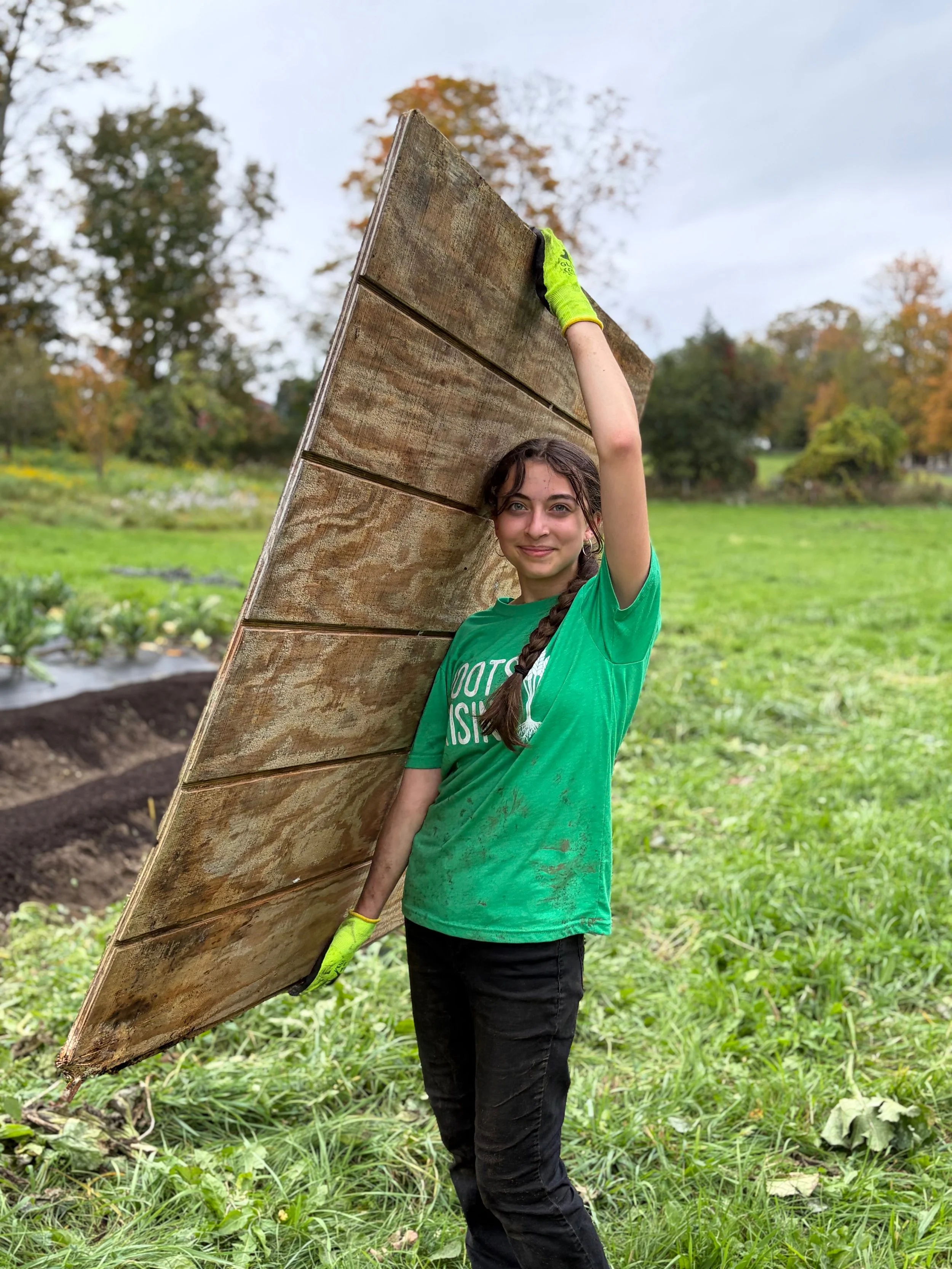 Roots Rising student in a green work glove holding up a plywood sheet in the field, autumn trees behind her