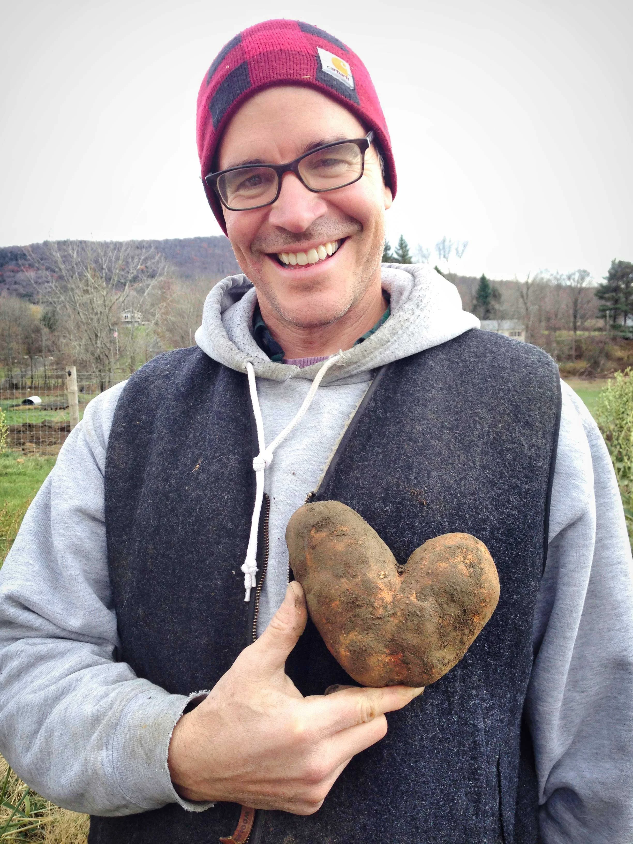 A man in a red plaid beanie and glasses holding a heart-shaped potato