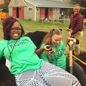 Two Roots Rising youth program participants laughing in a wheelbarrow while Jim Schultz shovels compost behind them at Red Shirt Farm