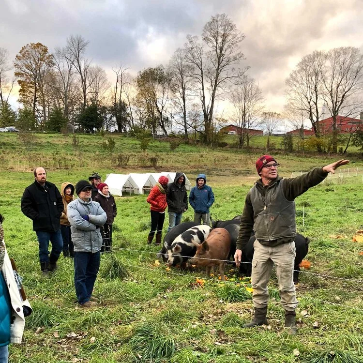 Jim Schultz leading a farm tour at Red Shirt Farm, gesturing across the pasture while visitors observe pigs on pasture in the background