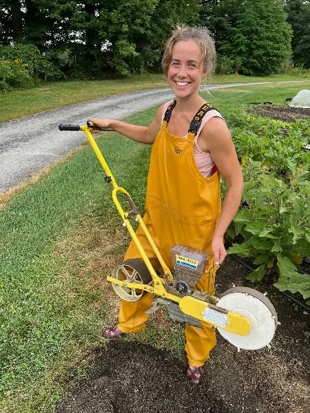 A smiling farmer in yellow overalls with a push seeder beside a gravel road and vegetable rows