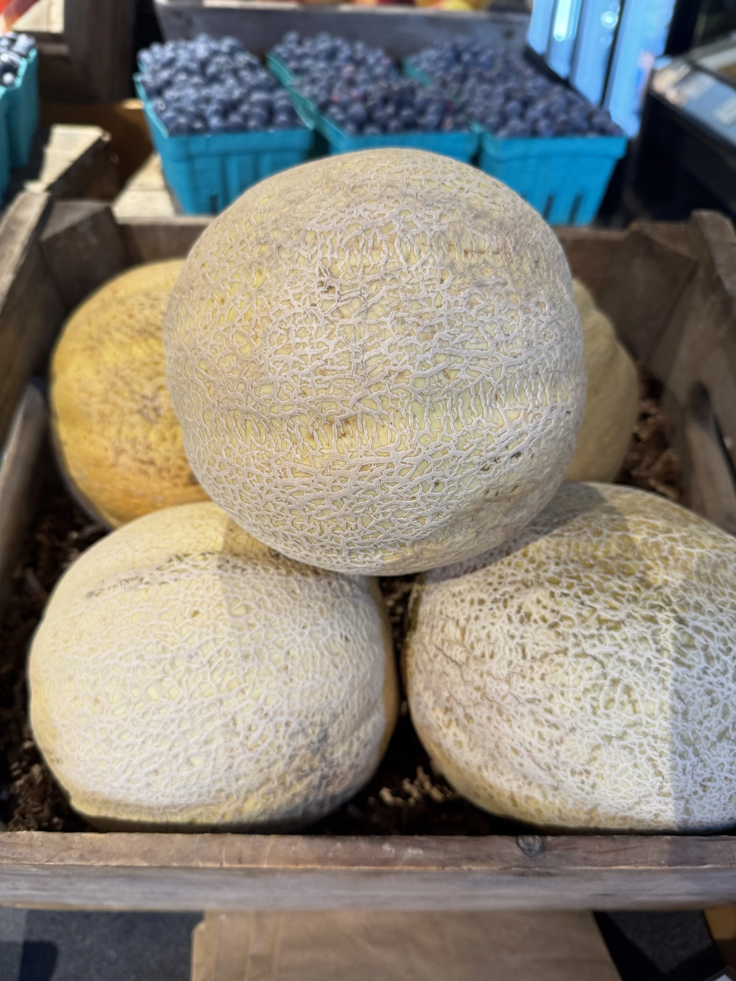 A cantaloupe in a wooden crate with cartons of blueberries behind it