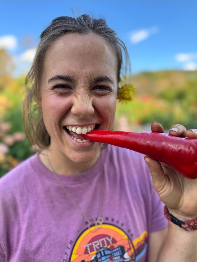 Sarah biting into a red Carmen pepper straight off the plant, with a yellow flower tucked behind her ear