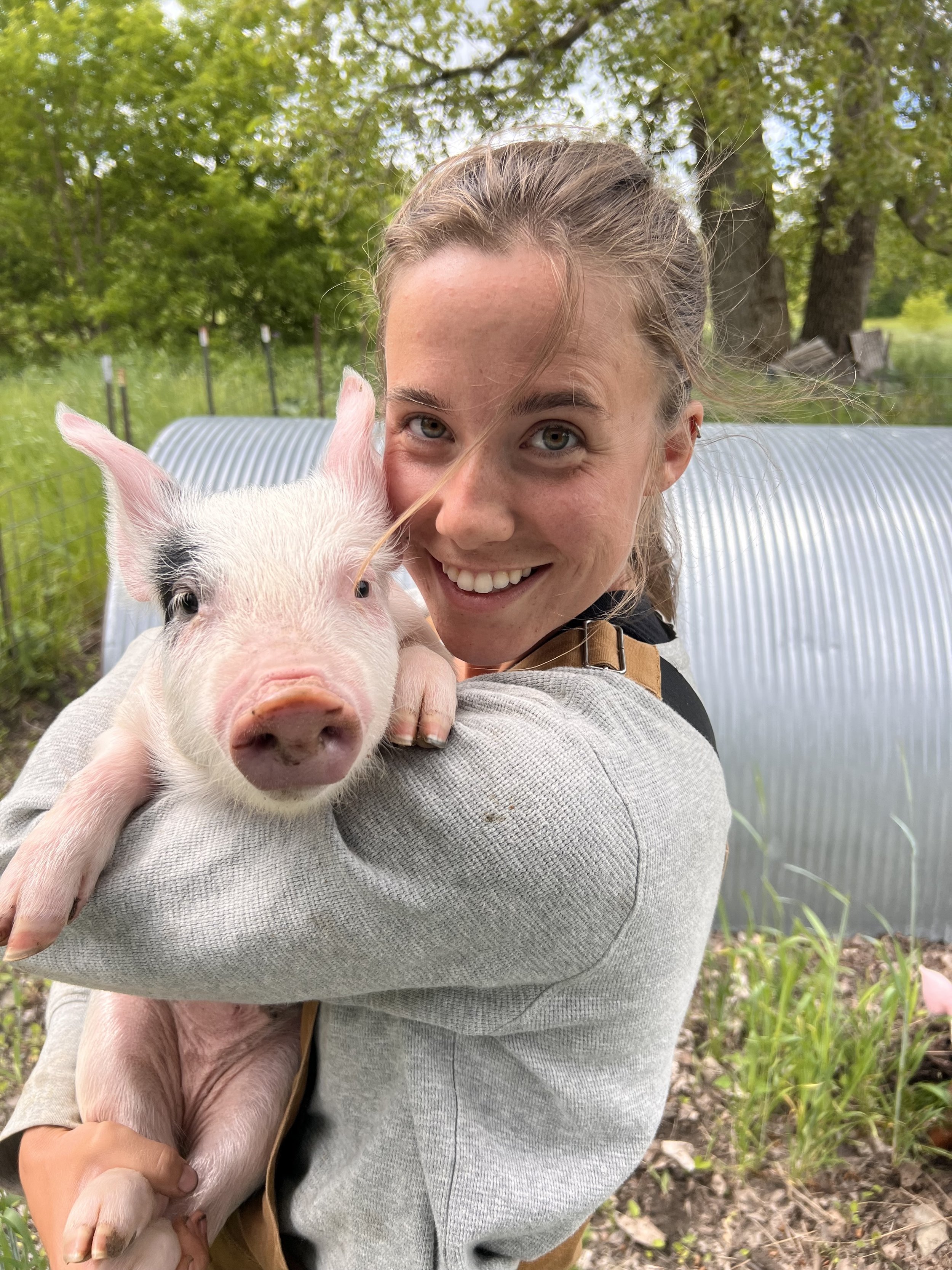 A woman holding a pink piglet against her cheek in front of a pasture hut