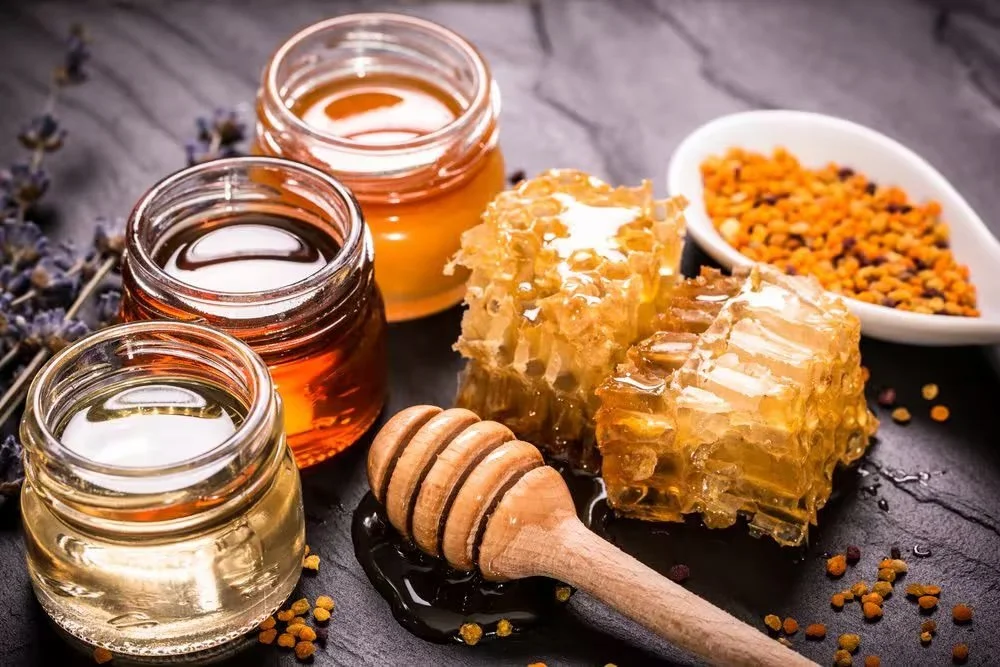 Jars of honey beside honeycomb and a bowl of bee pollen