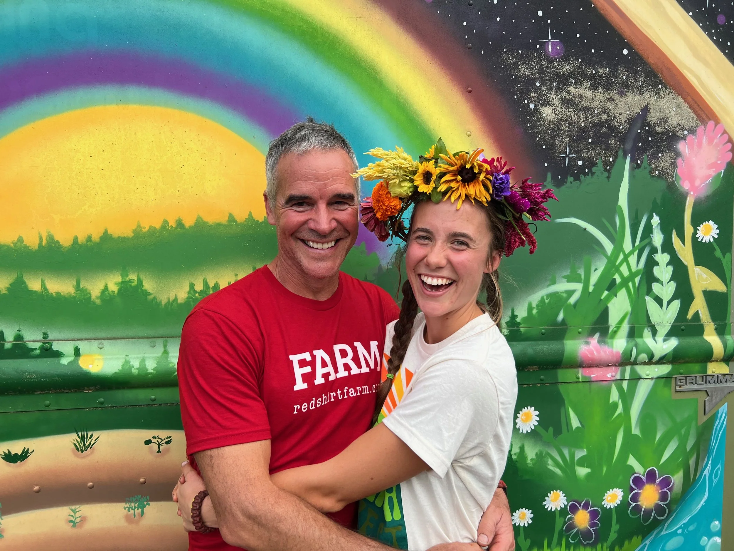 A man in a red farm shirt and a woman wearing a flower crown hugging in front of a rainbow mural
