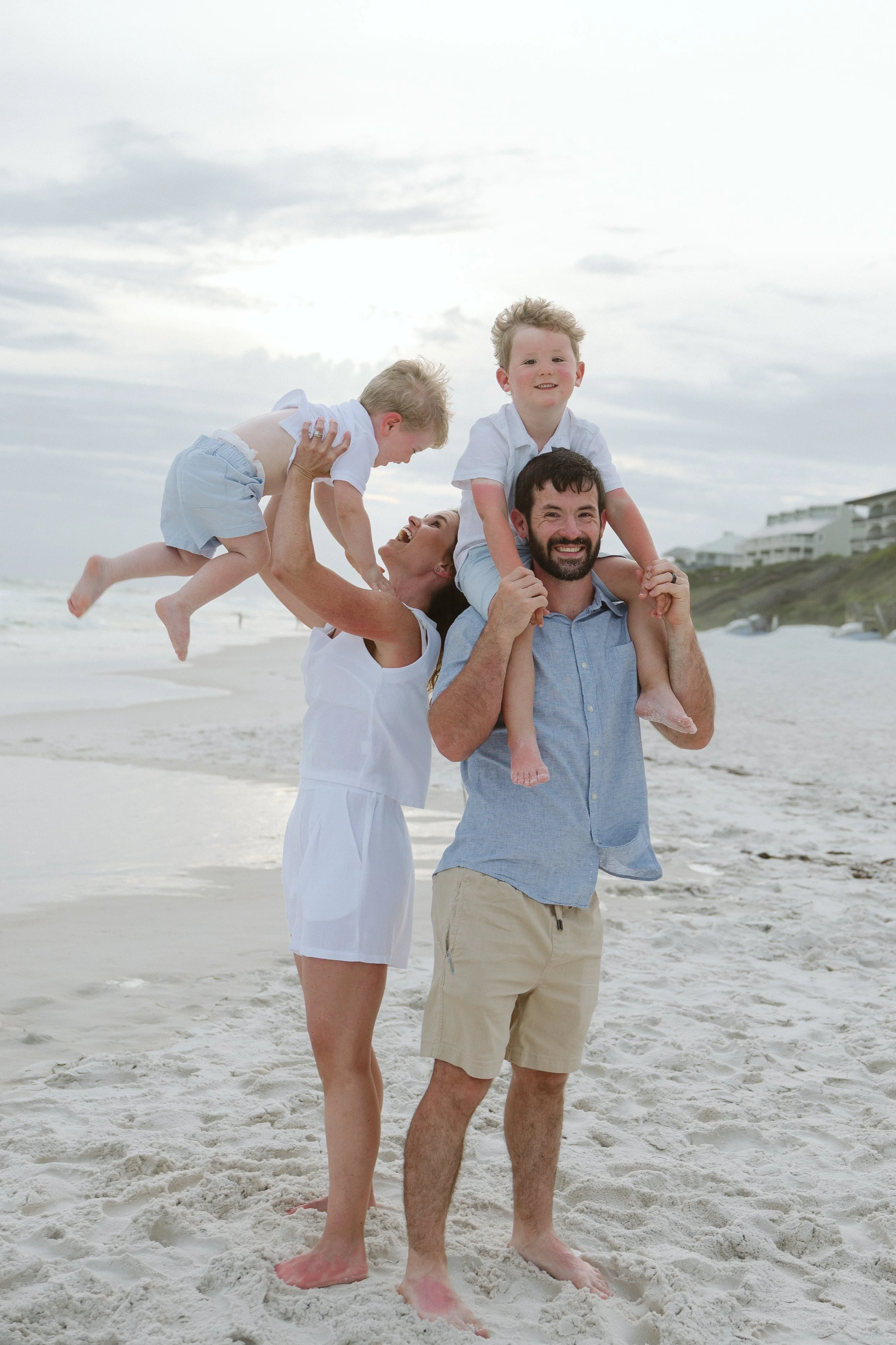 A family of four at the beach, with the father holding their young son on his shoulders and the mother holding their other young son, all smiling and enjoying the day.