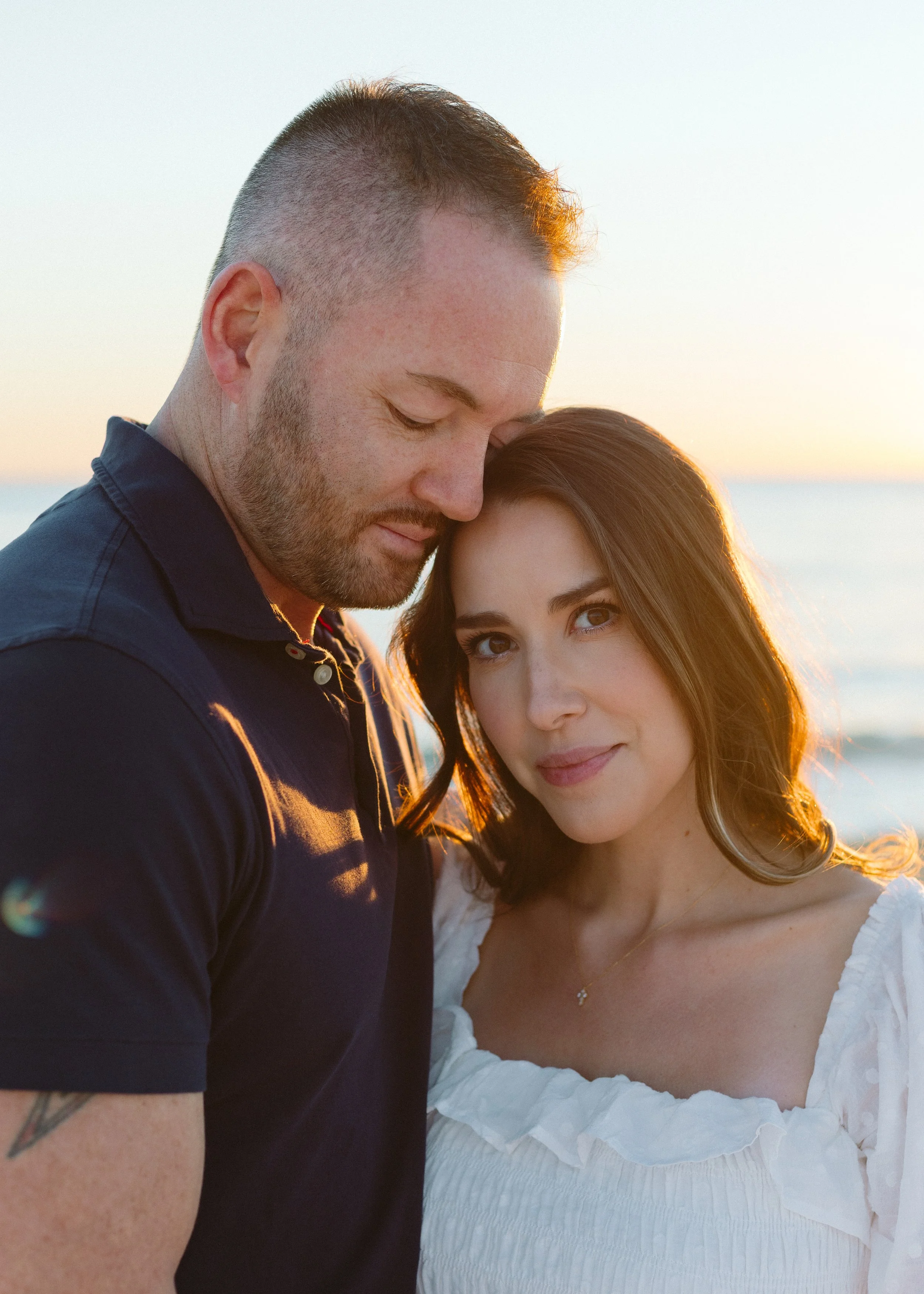 A couple standing close together at the beach during sunset, with the man resting his forehead on the woman's head and both looking at the camera.