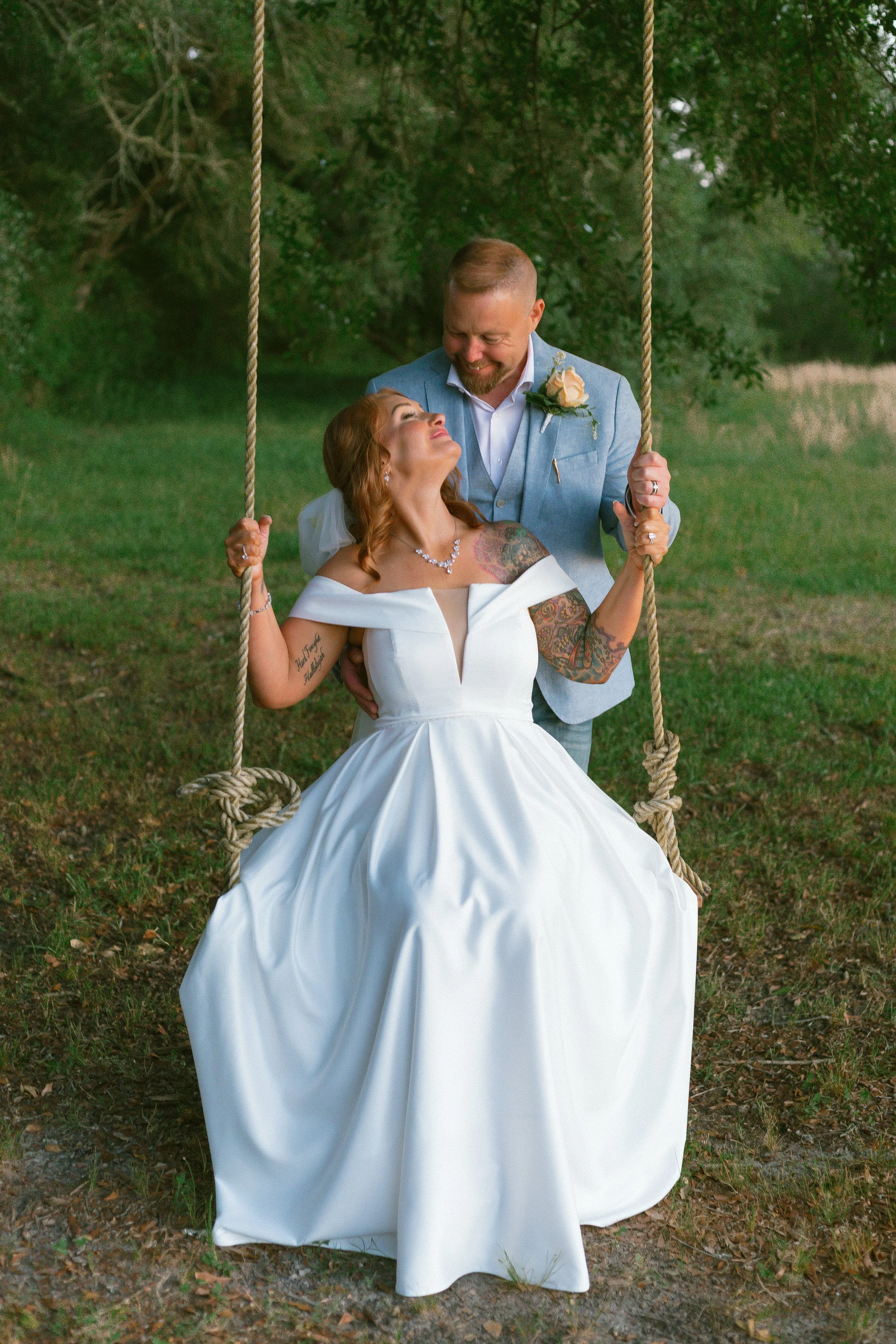 A bride and groom sharing a joyful moment on a swing outdoors. The bride is seated on the swing, wearing a white wedding dress with tattoos on her arms, and the groom is standing behind her, holding the ropes. The groom is dressed in a light blue suit with a white shirt and a boutonnière. They are smiling and looking at each other, surrounded by green trees and grass.