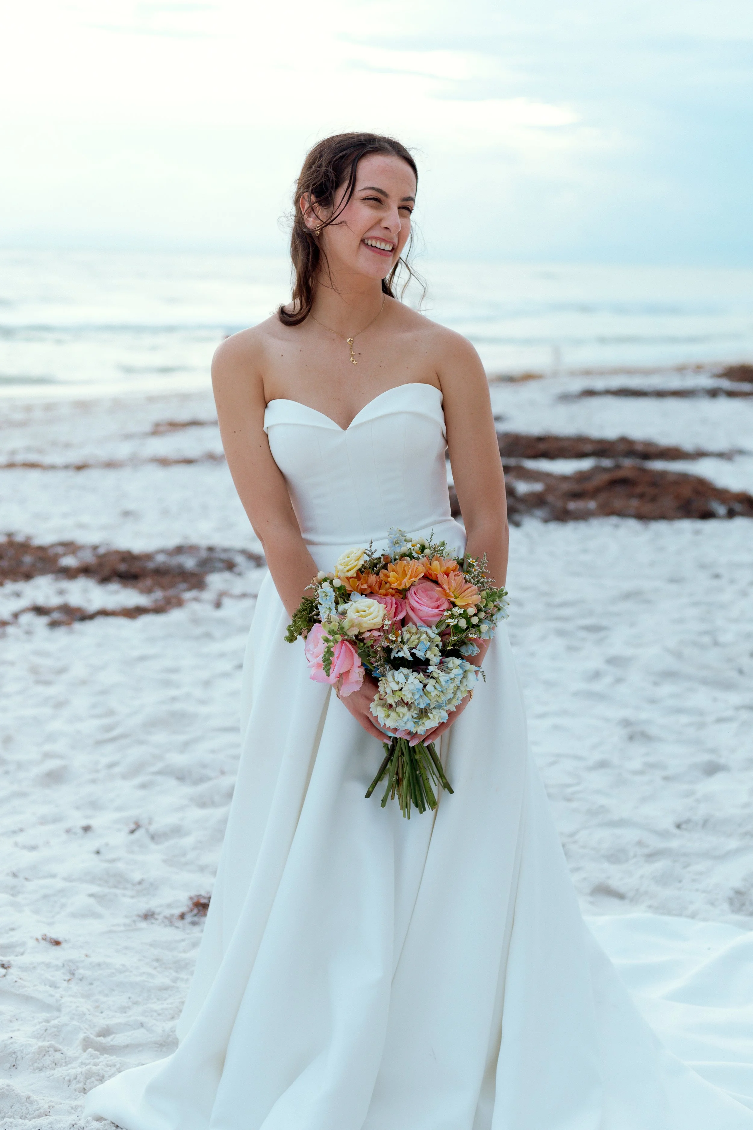 A woman in a white strapless wedding dress holding a colorful bouquet of flowers, standing on a sandy beach with ocean waves in the background, smiling and winking.