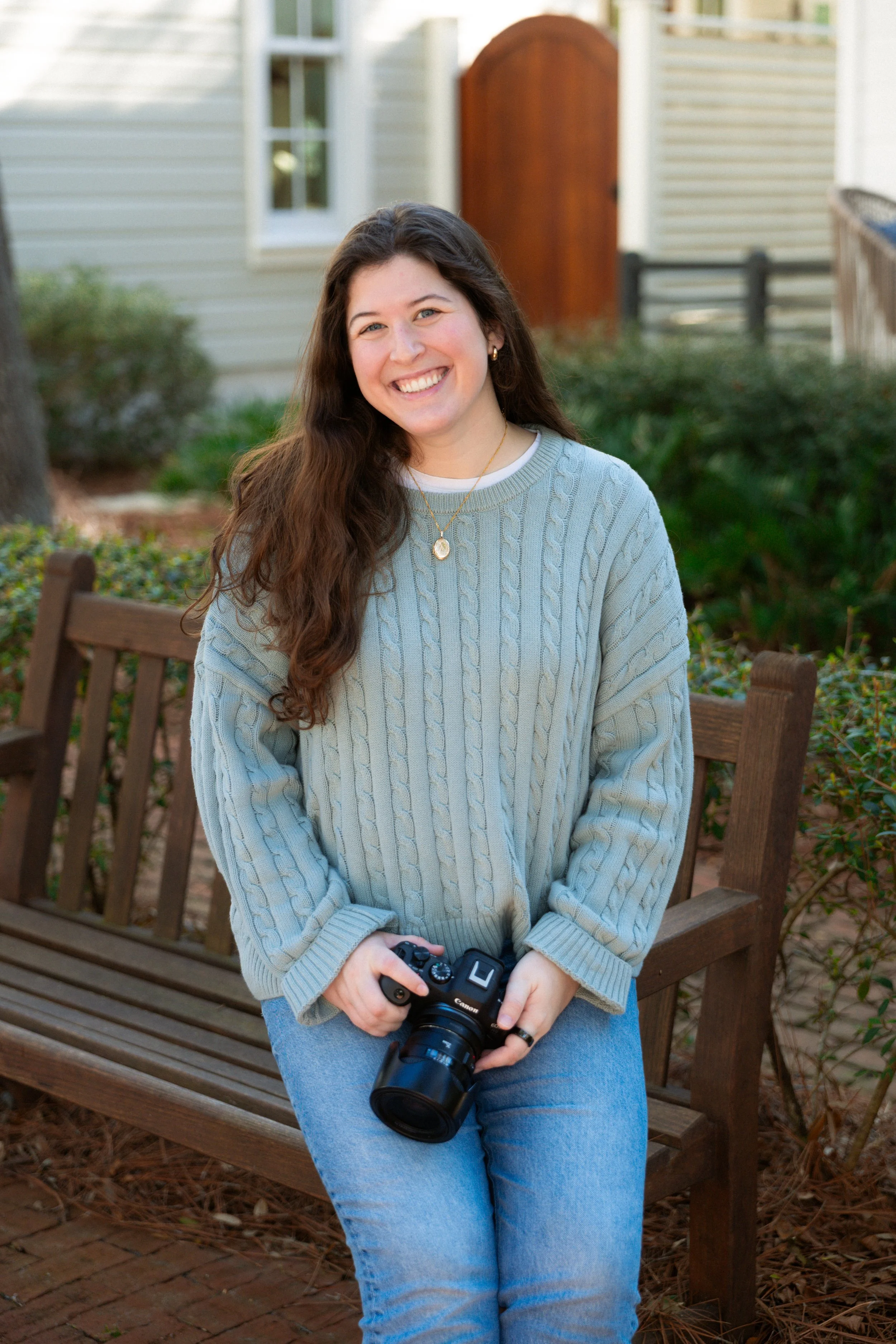 Smiling woman with long brown hair sitting on a wooden bench outdoors, holding a camera, wearing a light gray cable-knit sweater and blue jeans.