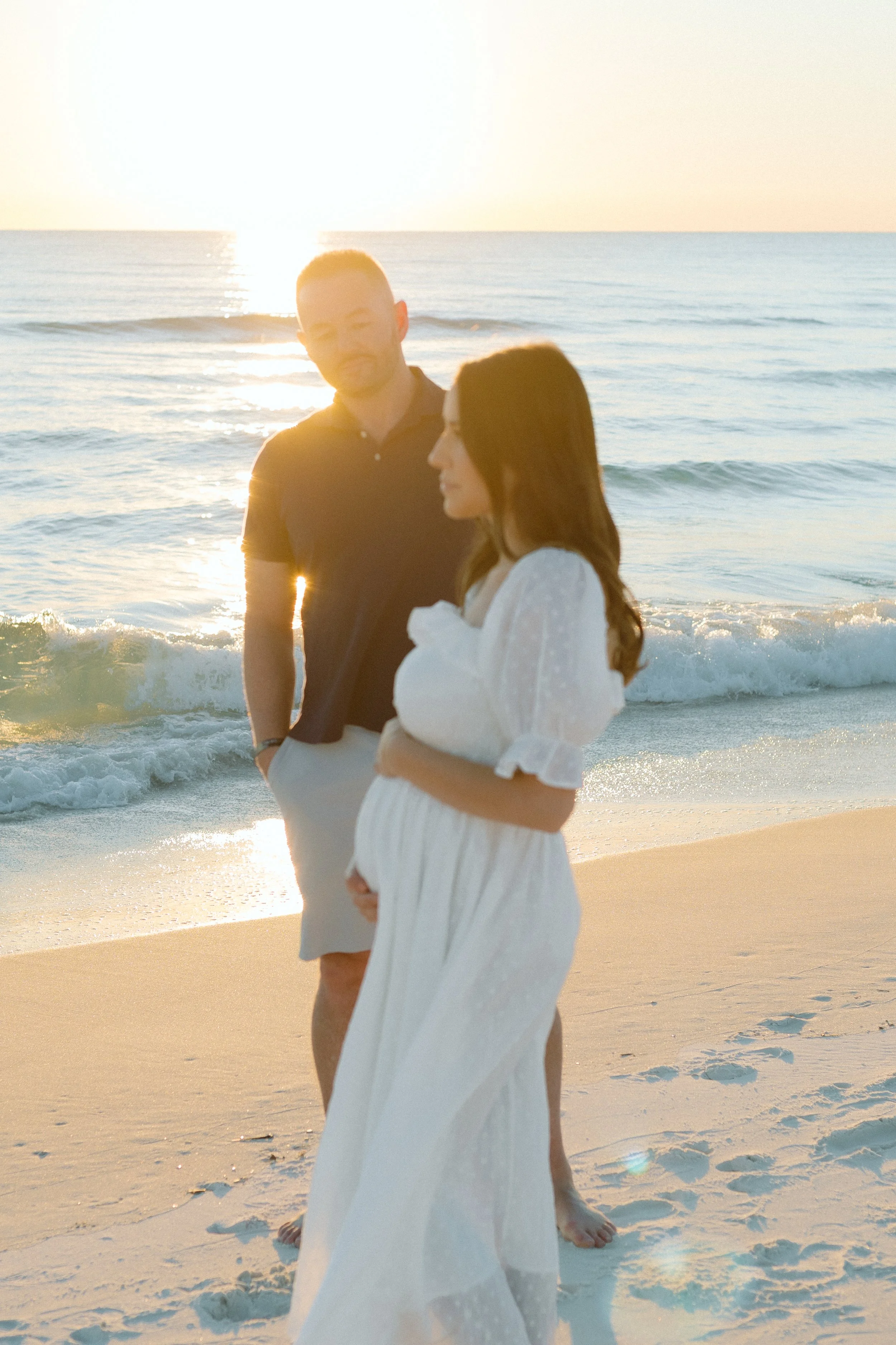 A man and woman standing on the beach during sunset near the ocean, with the sun low on the horizon and waves in the background. The woman is wearing a white dress, and the man is dressed in a dark shirt and light shorts.