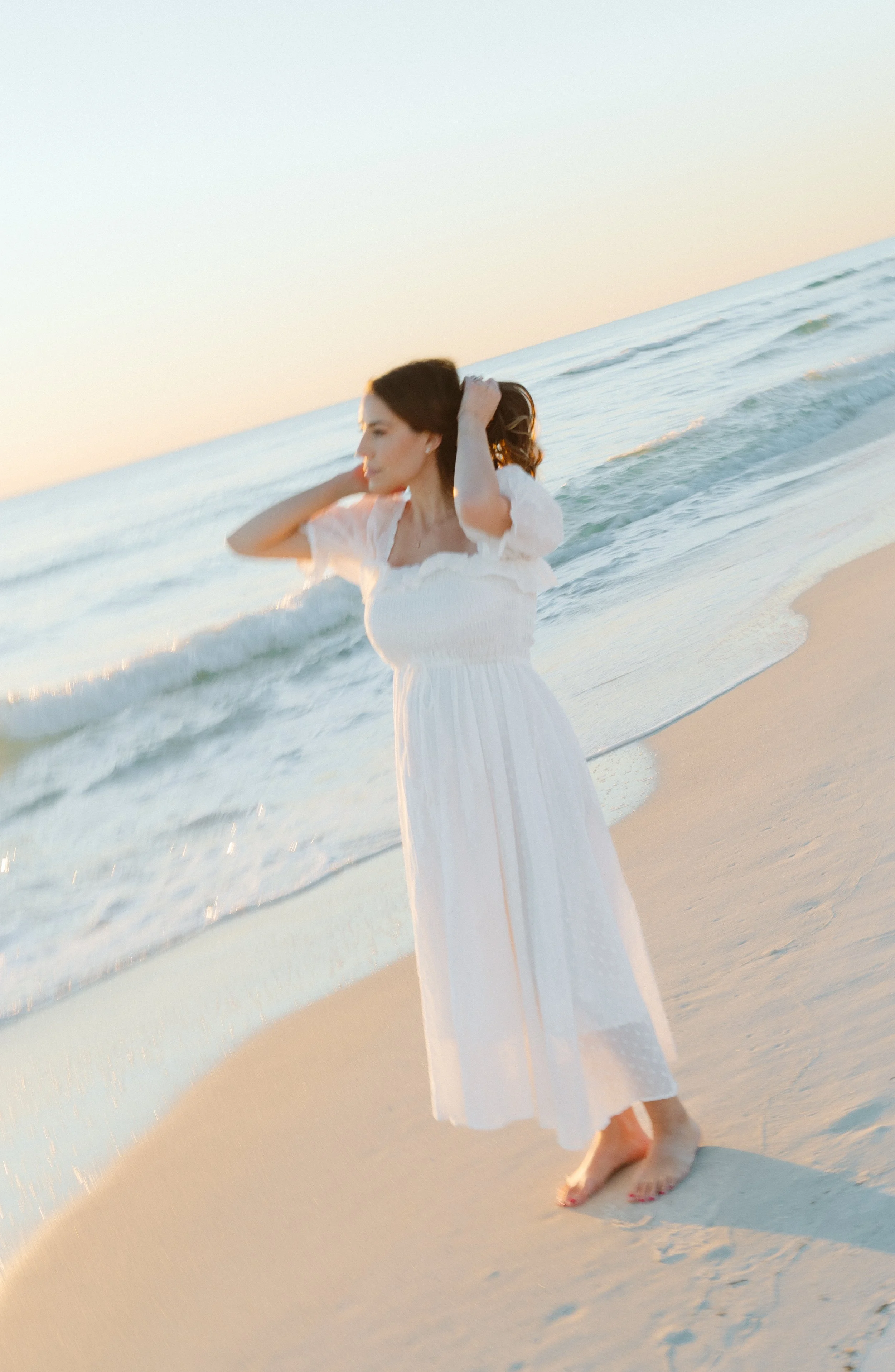 A woman in a white dress standing barefoot on the beach near the water at sunset, with her arms raised and hands in her hair.