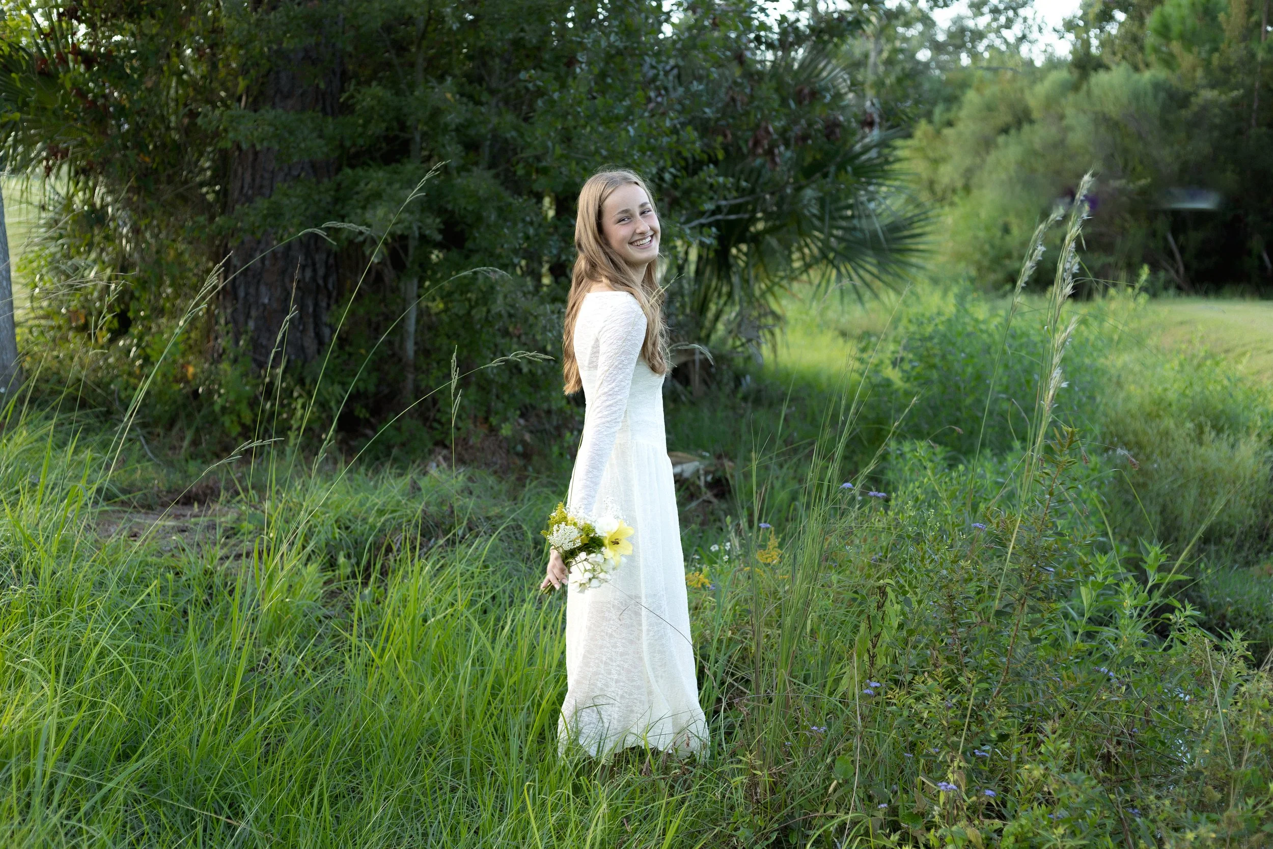 A young woman in a white dress outdoors, holding a small bouquet of flowers, standing in tall grass near trees, smiling.