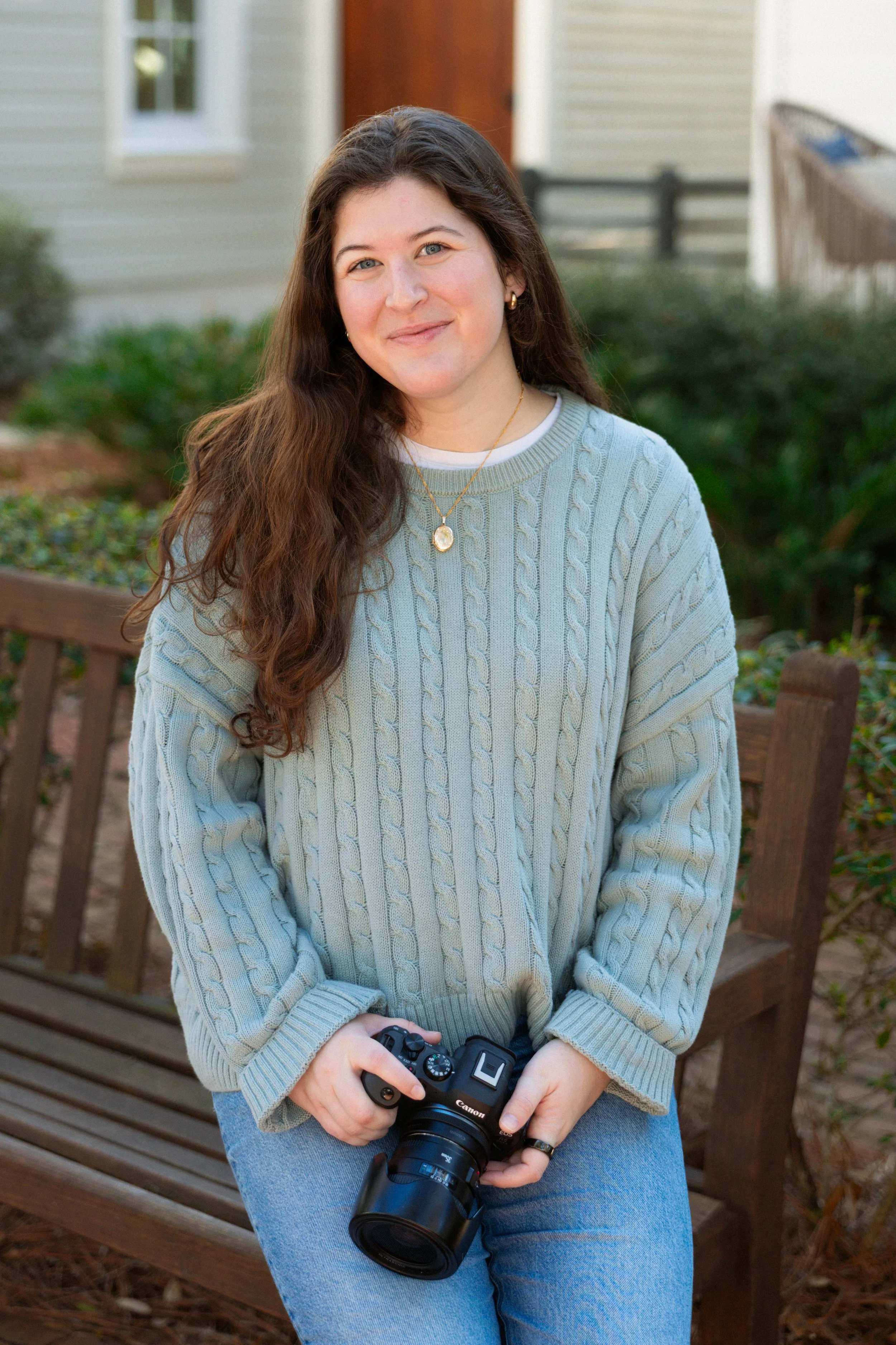 A young woman with long brown hair, wearing a light blue cable-knit sweater and jeans, holding a camera, sitting on a wooden bench outdoors, smiling at the camera.