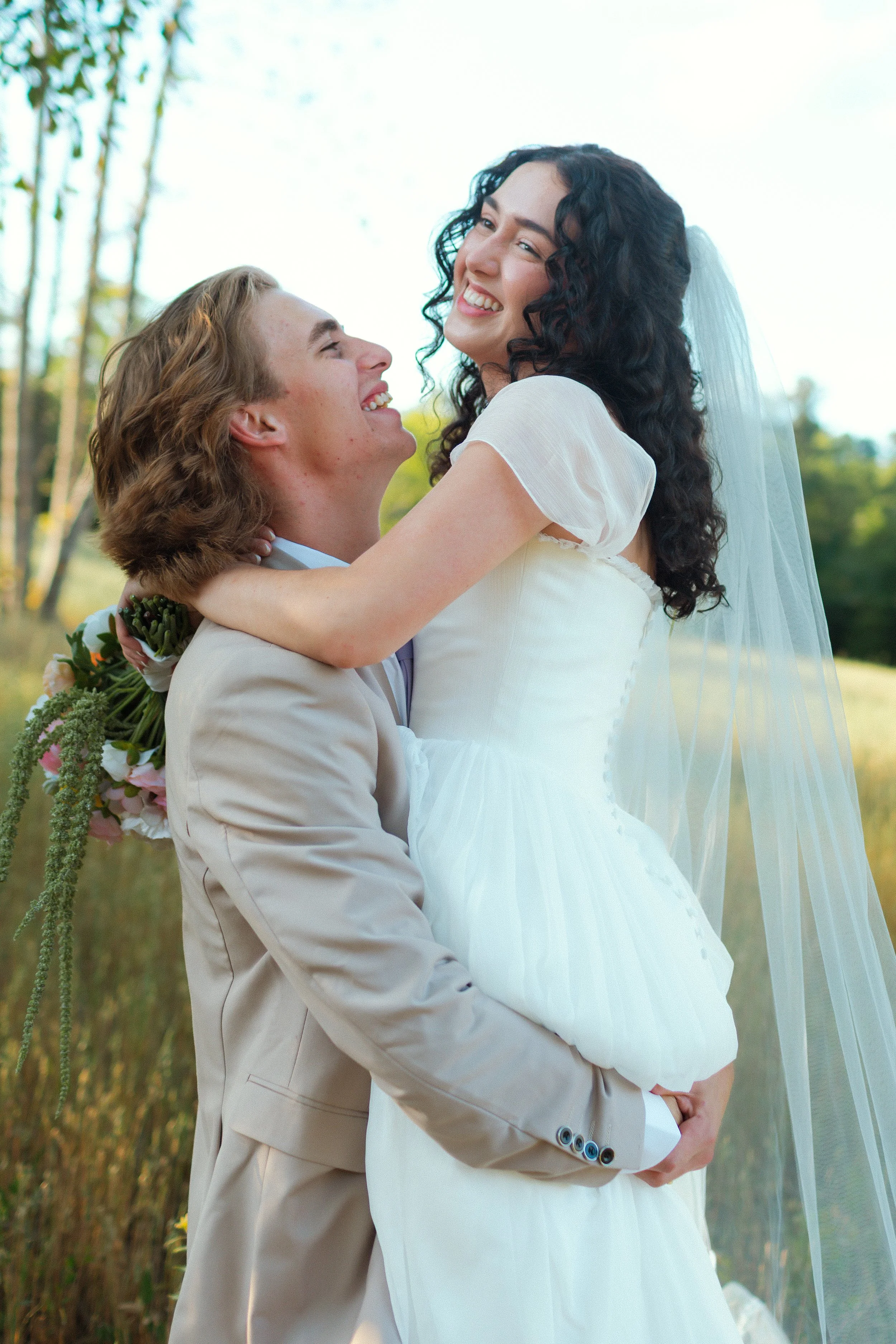 A smiling couple, the woman in a white wedding dress and veil, and the man in a beige suit, embracing outdoors during daytime.