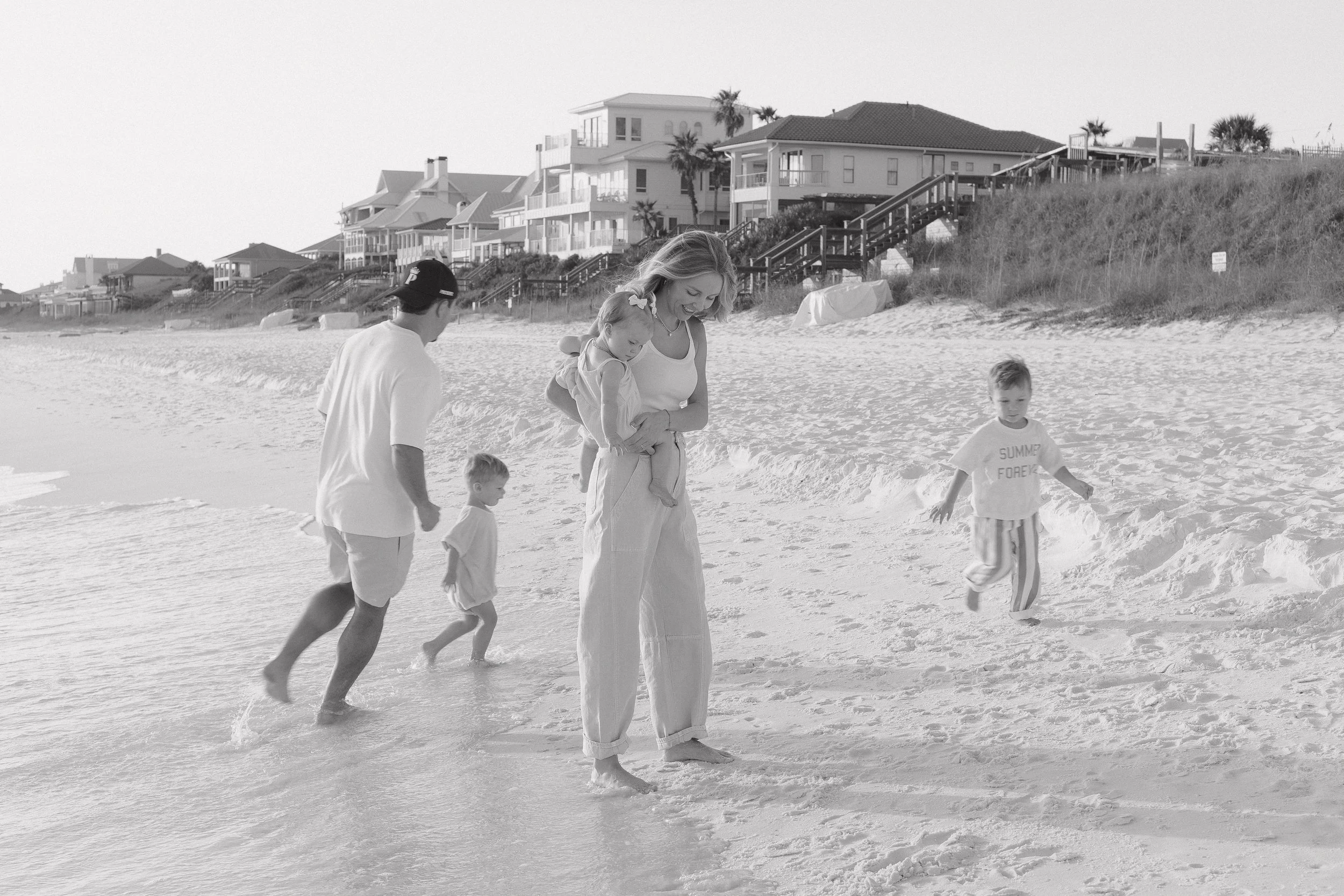 A family of five, including two children and two adults, walking along the beach near the water, with houses and palm trees in the background.