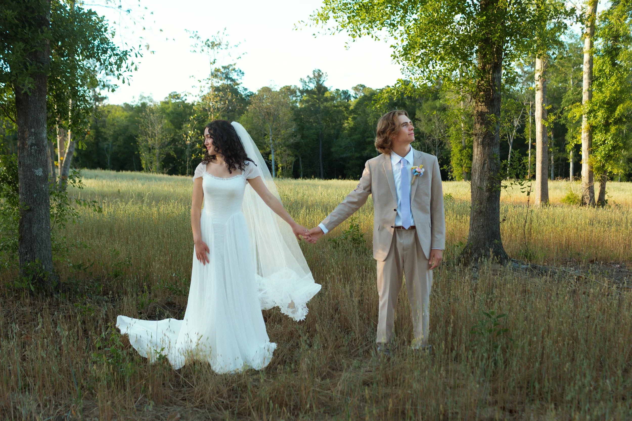 A bride and groom holding hands in a grassy field surrounded by trees, with the bride in a white wedding dress and veil, and the groom in a beige suit with a boutonniere, during a sunny day.