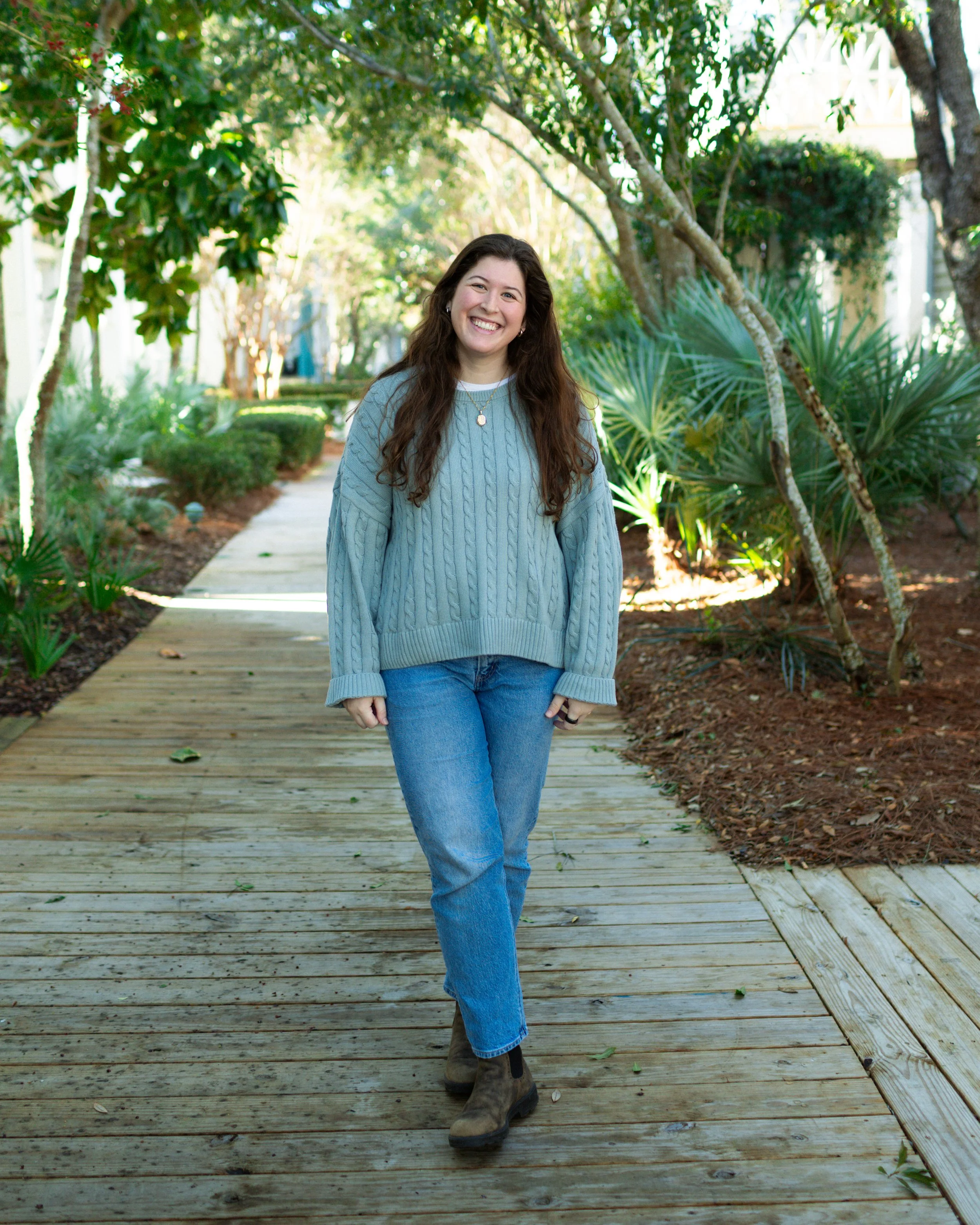 A young woman with long brown hair smiling while walking on a wooden path surrounded by green trees and plants