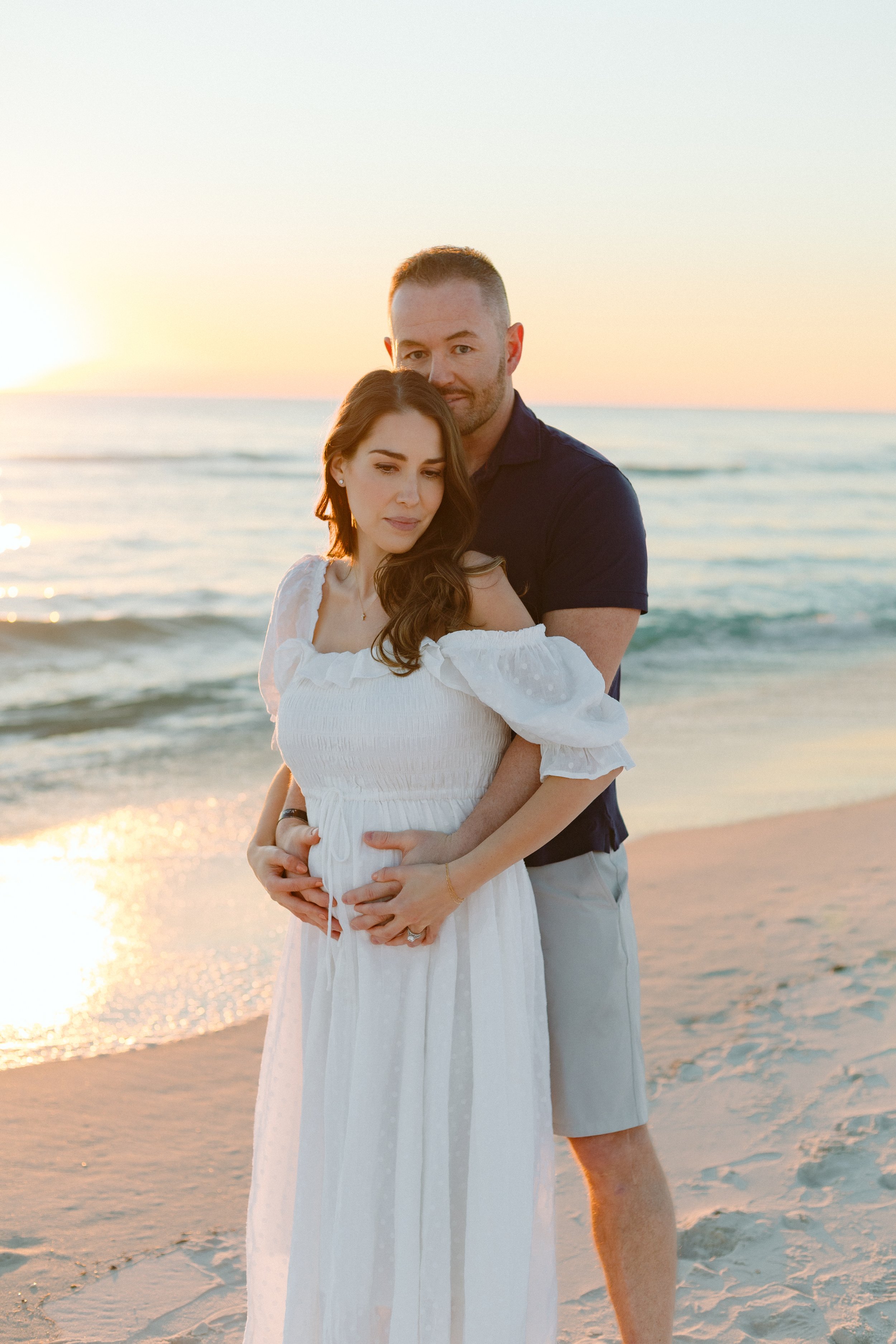 A couple standing on the beach during sunset, with the ocean in the background. The woman is wearing a white dress, and the man is wearing a navy shirt and shorts. They are holding each other, looking contemplative.
