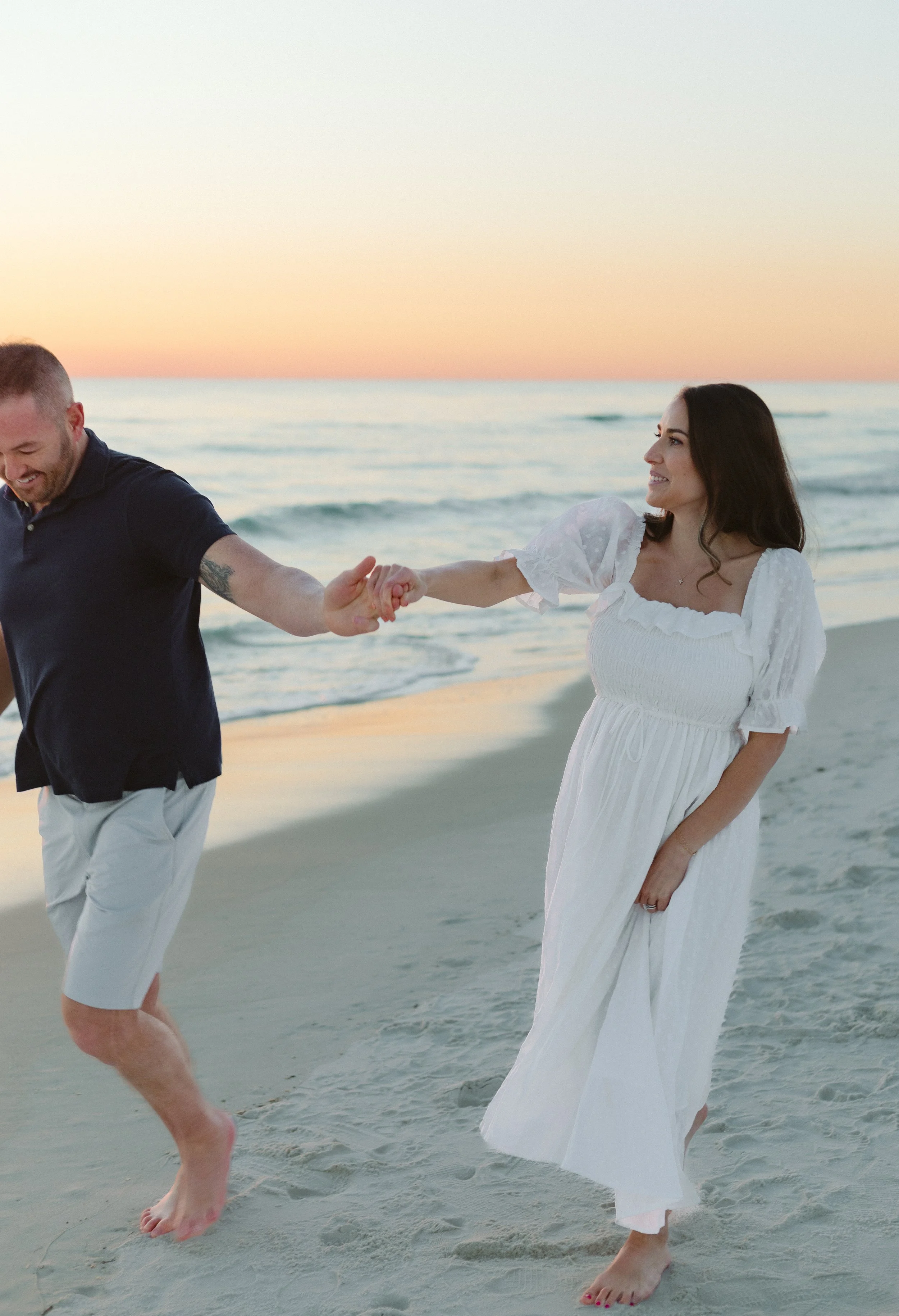 A couple holding hands and playing on the beach during sunset, woman in a white dress and man in a dark shirt and light shorts.