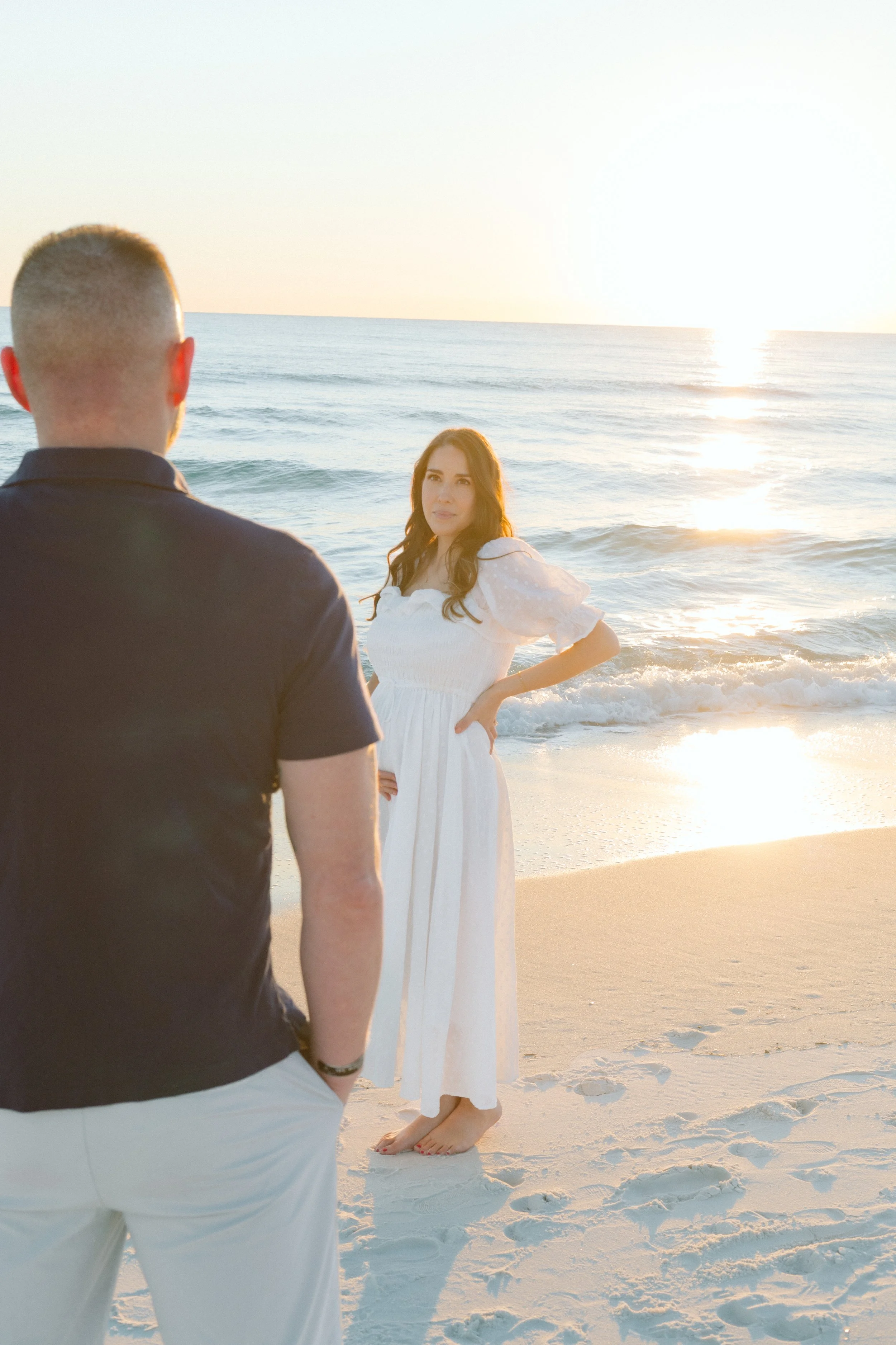 A man and woman on a beach during sunset, facing each other; the woman has long dark hair and wears a white dress, and the man has short hair, wearing a dark shirt and light-colored pants.