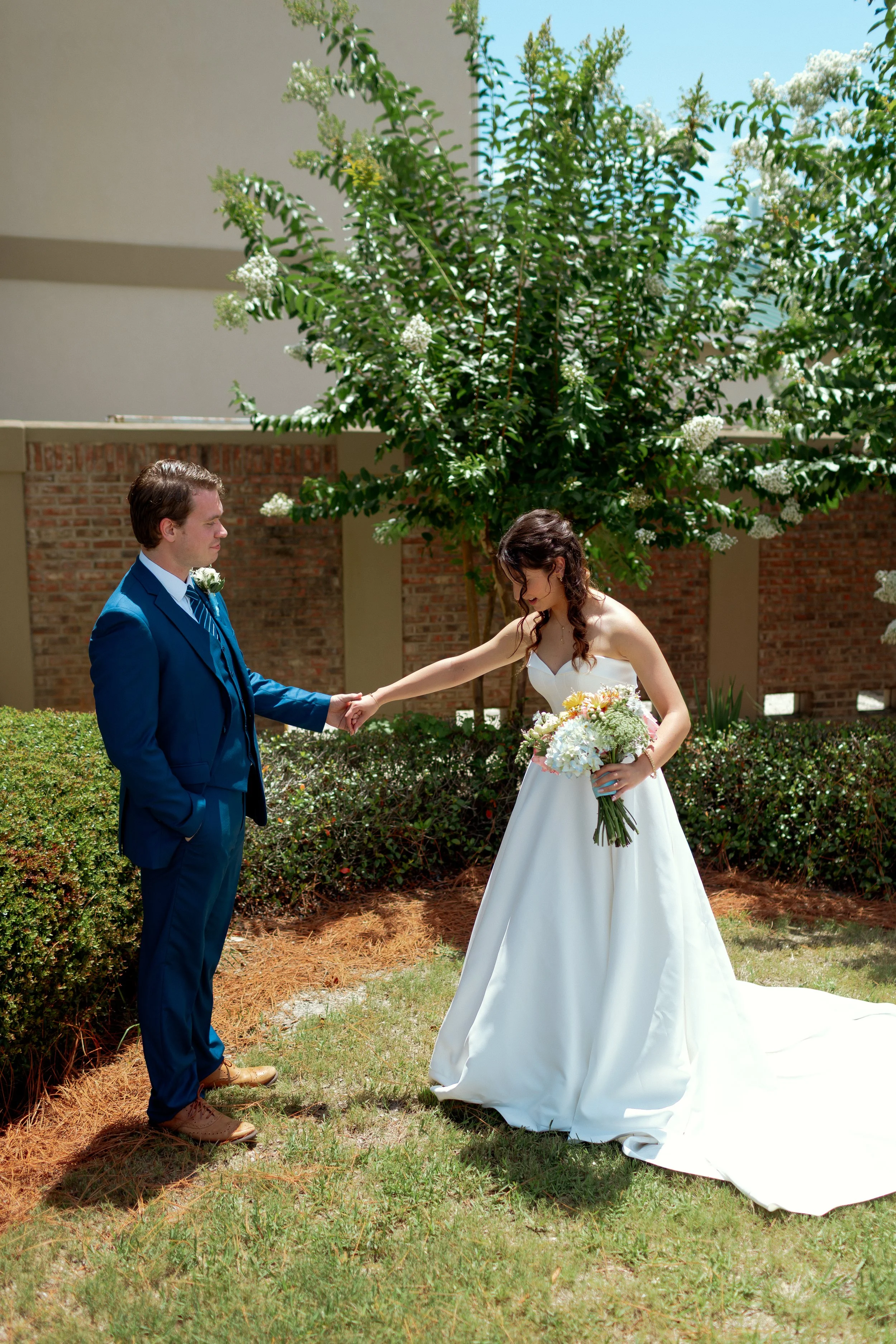 A bride and groom holding hands outdoors during a wedding ceremony. The groom in a blue suit, and the bride in a white wedding dress holding a bouquet of flowers. There is a large green tree, bushes, and a brick wall in the background.