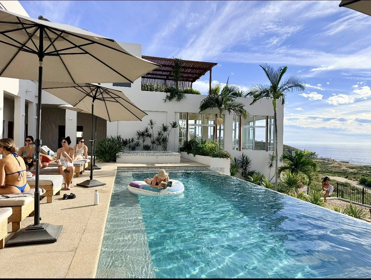 a group of girls hang out on chaises by a pool in Mexico, while one girl floats on a floatie in the pool. It's a beautiful day with the ocean in the background.