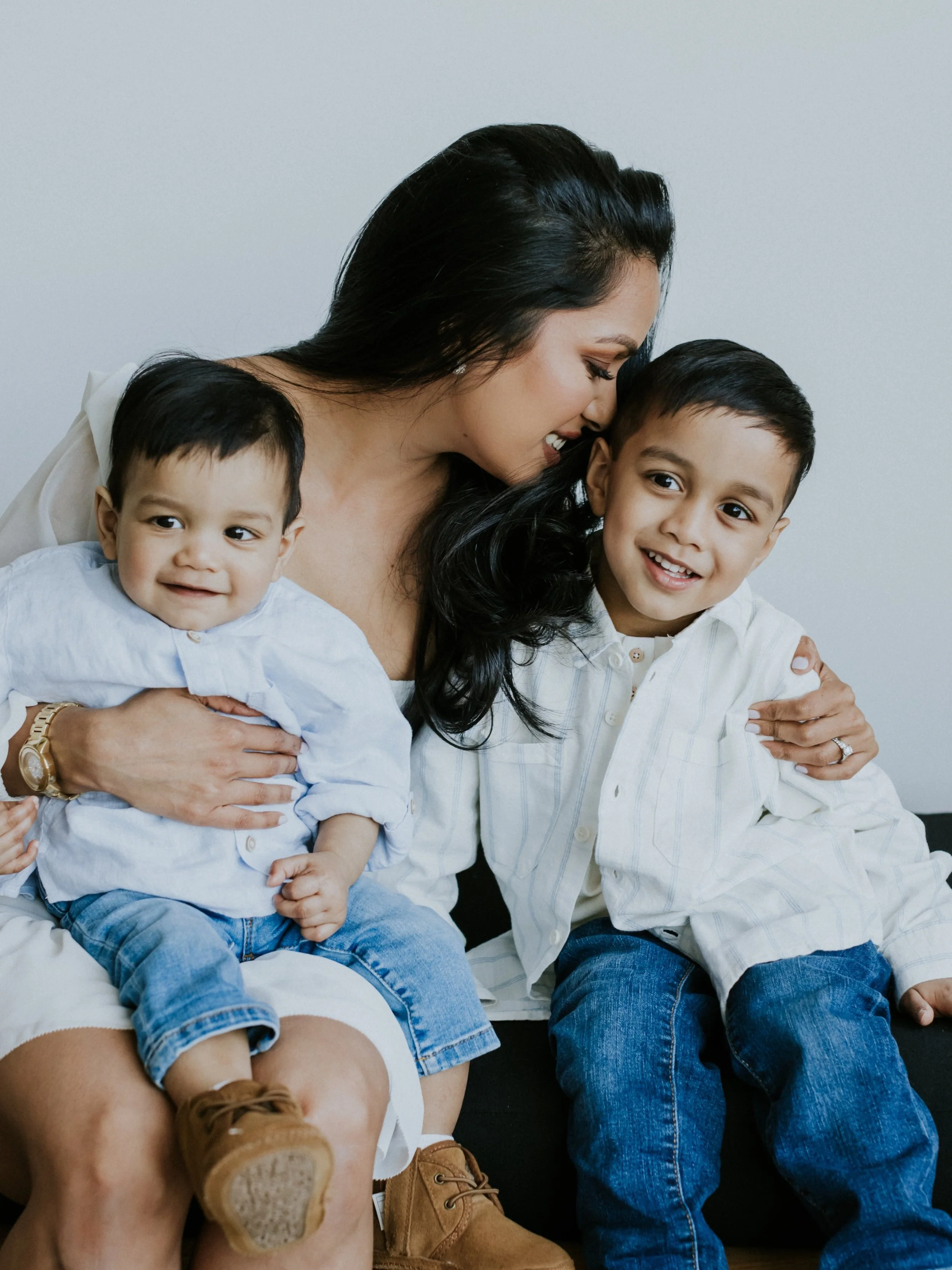 A woman sitting on a couch with two children, smiling and wearing casual attire.