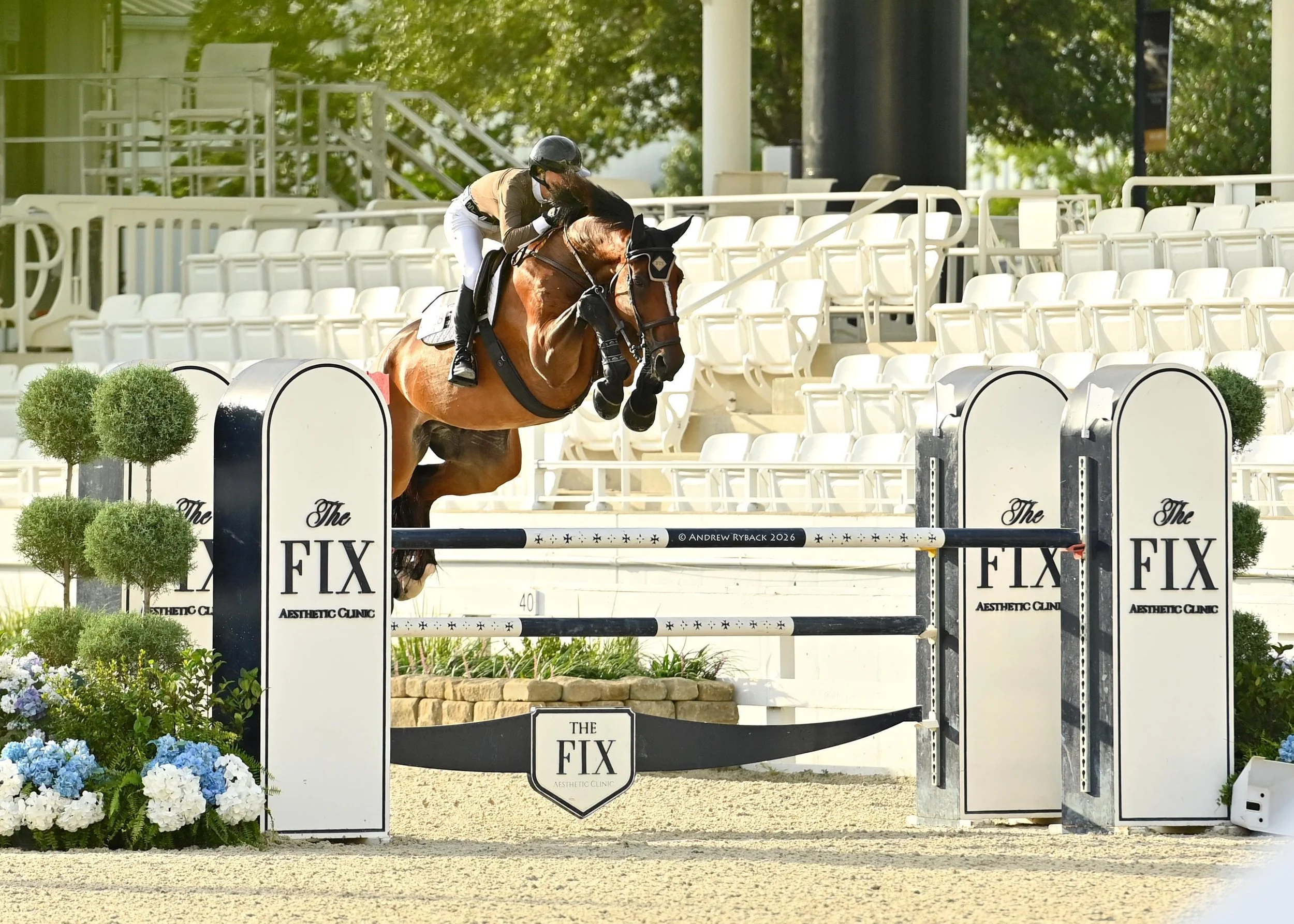 A person wearing riding gear, including a helmet and gloves, riding a brown horse over an obstacle at an equestrian event, with empty white stadium seats in the background.