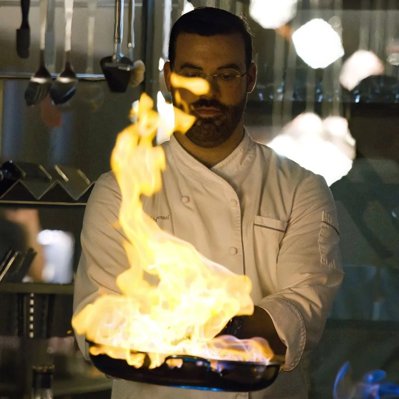 A chef wearing glasses and a white chef's coat is creating a flame show over a black pan in a professional kitchen.