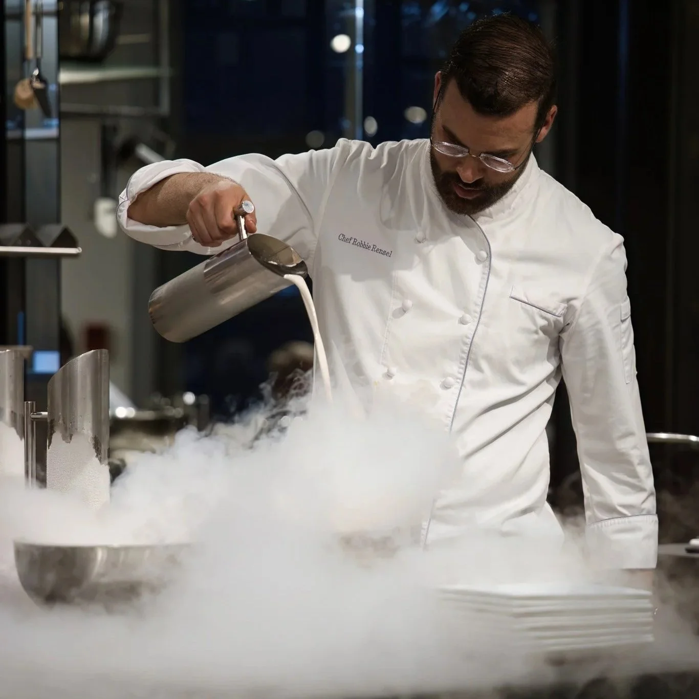 Chef pouring liquid into bowl with smoke or vapor rising from it, in a commercial kitchen.