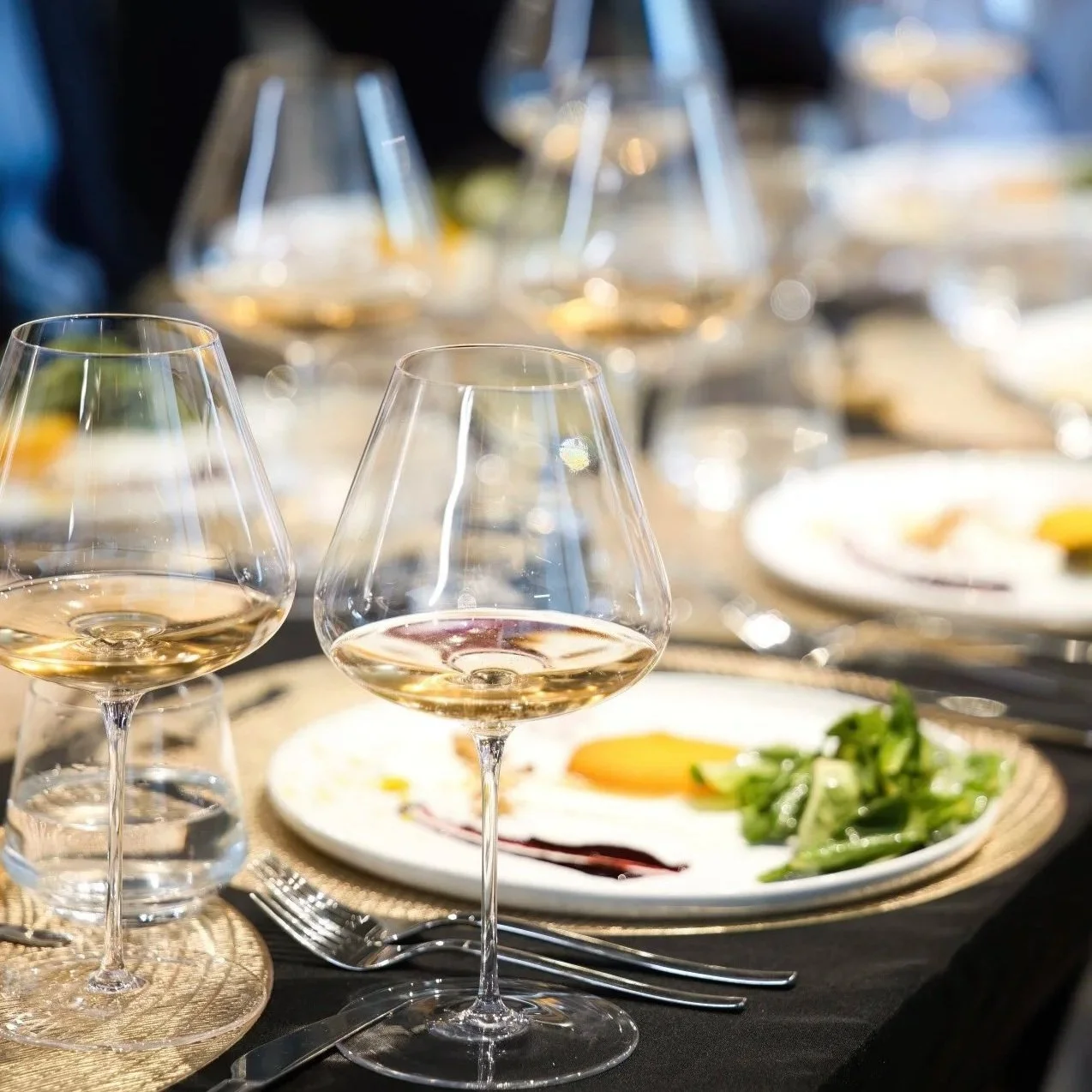 Several glasses of white wine on a dining table with plates of food, including salad and a sunny-side-up egg.