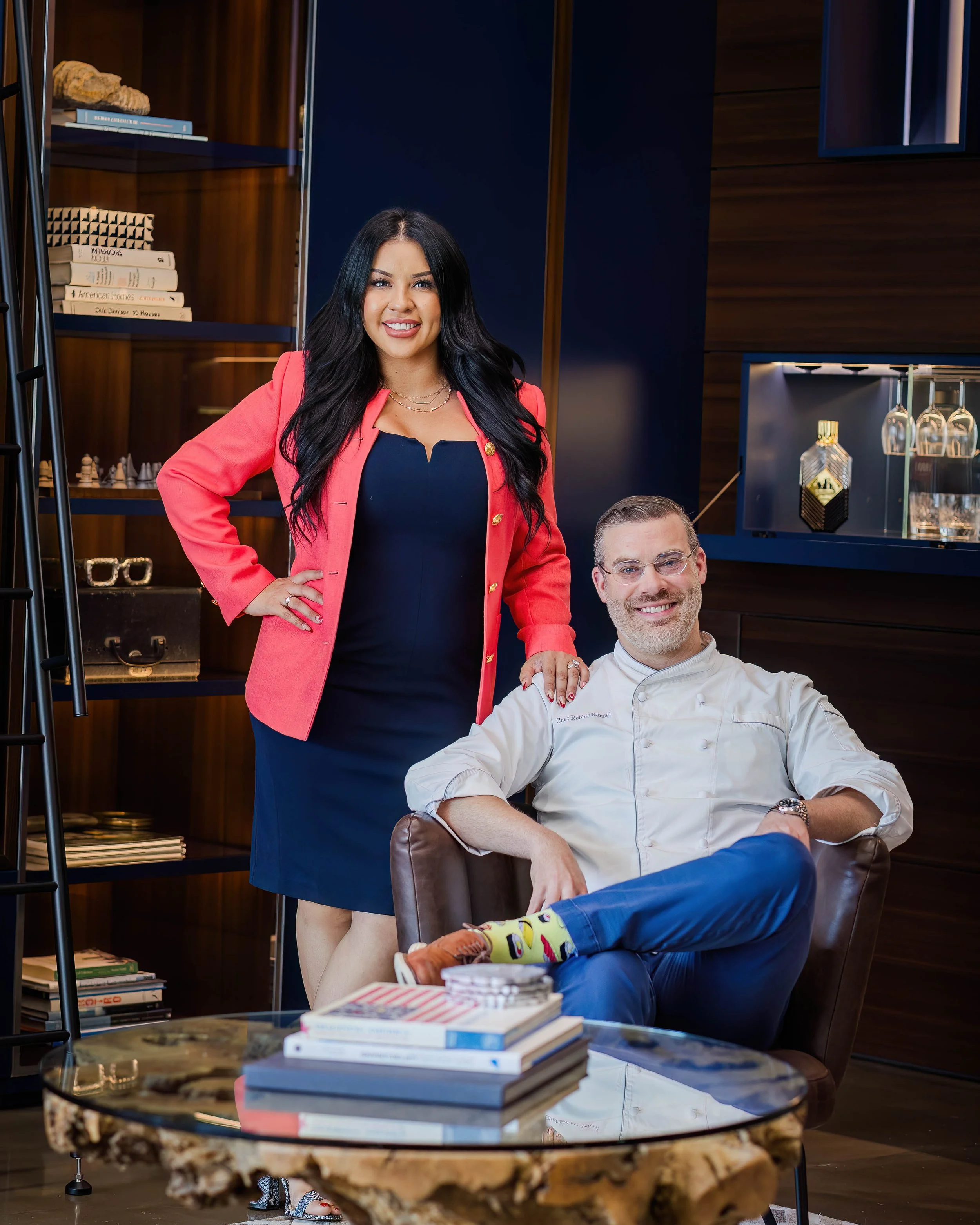 A woman in a pink blazer and navy dress standing next to a seated man in an office or lounge with bookshelves and decorative items in the background. She has long black hair and is smiling, resting her hand on the man's shoulder.