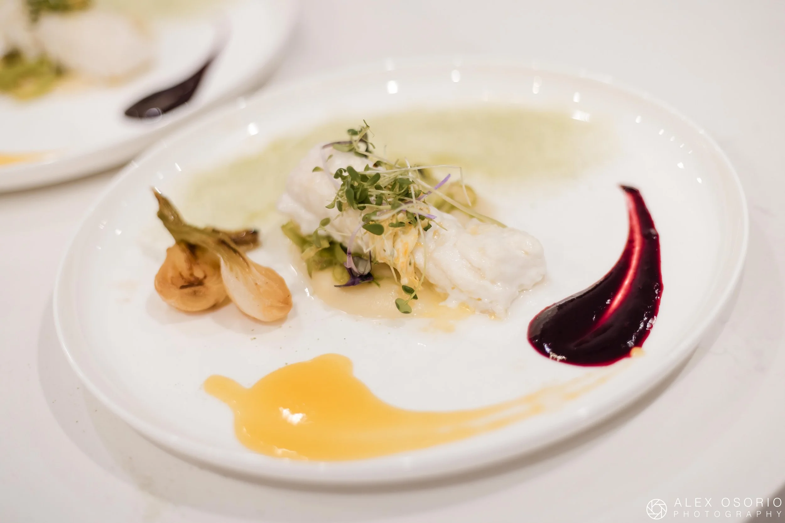 Plated fish course with vegetables, microgreens, and colorful sauces.