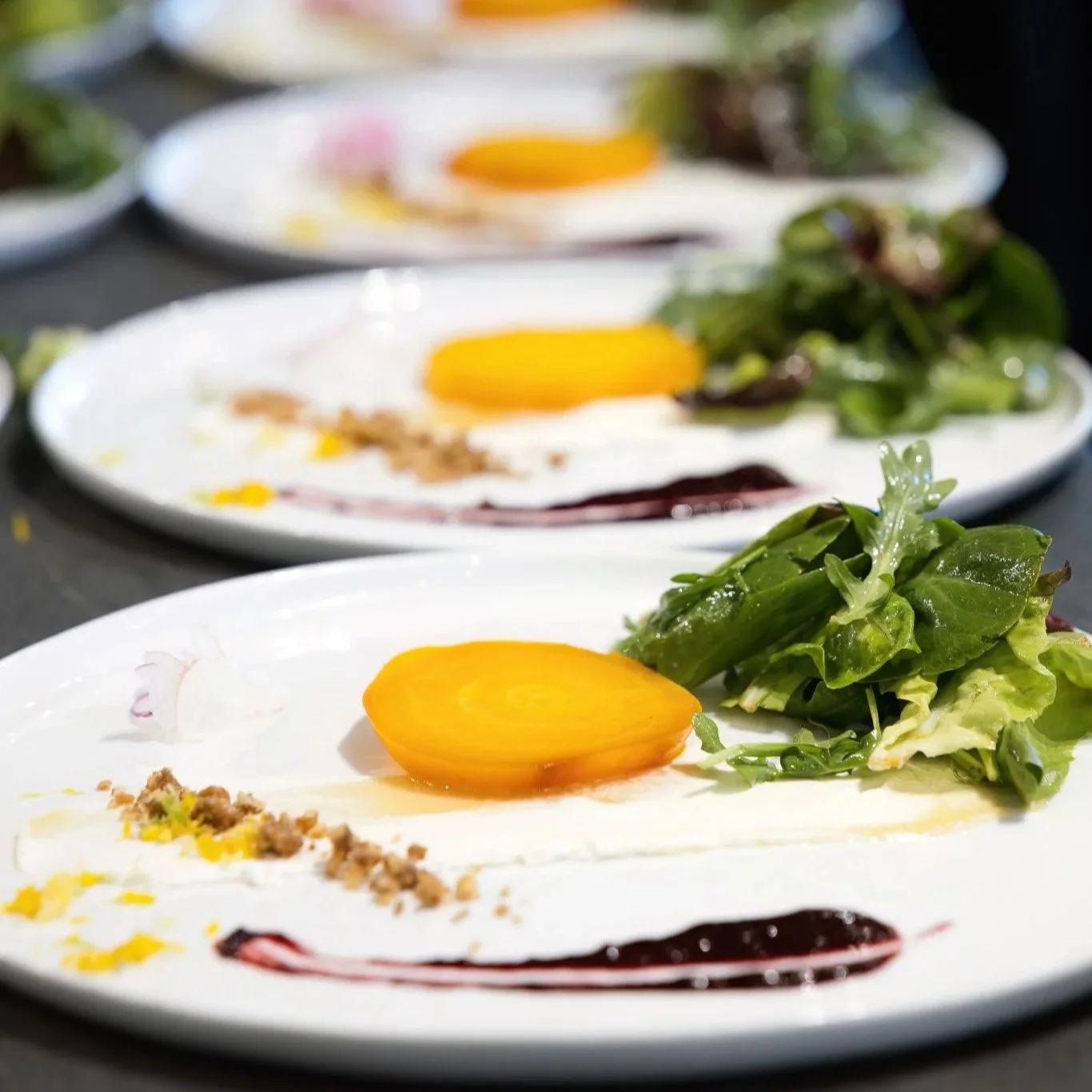 Close-up of plates with salad, yellow cherry tomato, and sauce, with blurred plates in background.