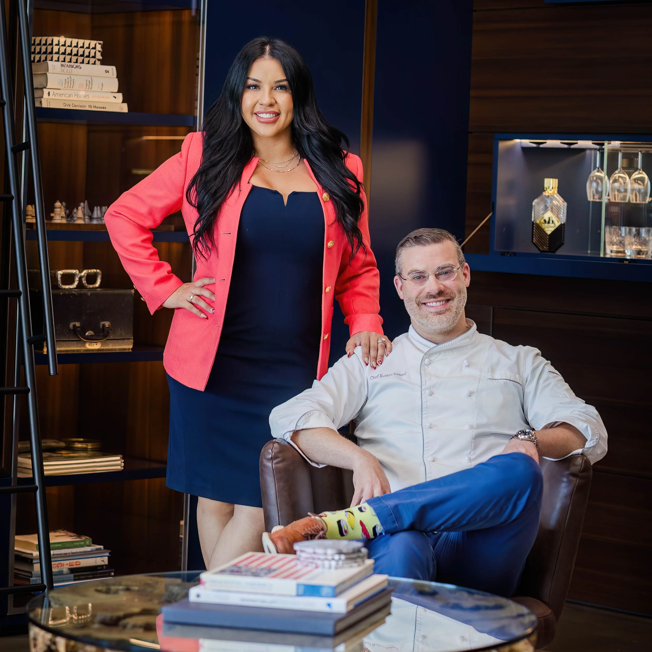 A woman with long black hair, wearing a coral blazer and navy dress, standing beside a man in a white chef's coat in a modern room with wooden shelves, books, and decorative objects.