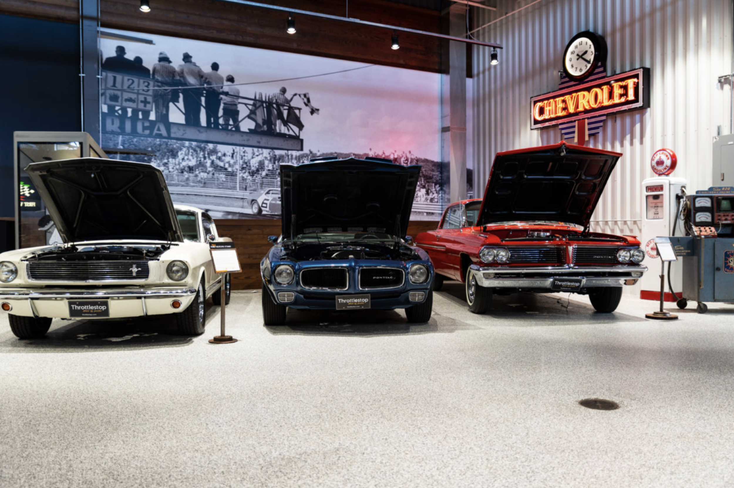 Classic car exhibition featuring a white Ford Mustang, a blue Pontiac, and a red Pontiac with hoods open, set in a retro-themed museum with vintage signage and gas station decor.