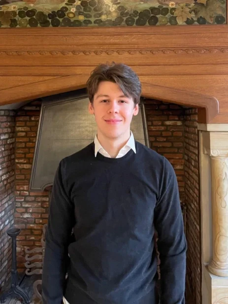 A smiling woman with curly hair wearing glasses, hoop earrings, layered necklaces, and a striped shirt in an indoor setting.