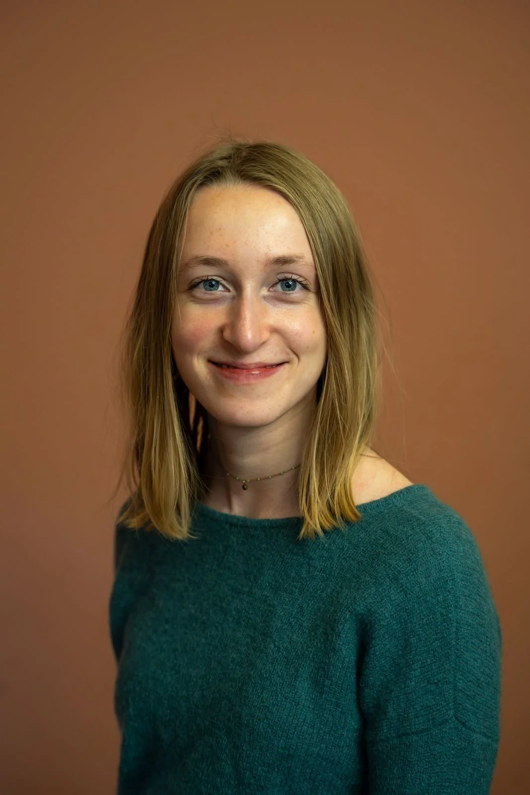 A young woman with short, dark, slightly tousled hair and blue eyes smiling outdoors, standing in front of an ornate, carved stone wall or building.