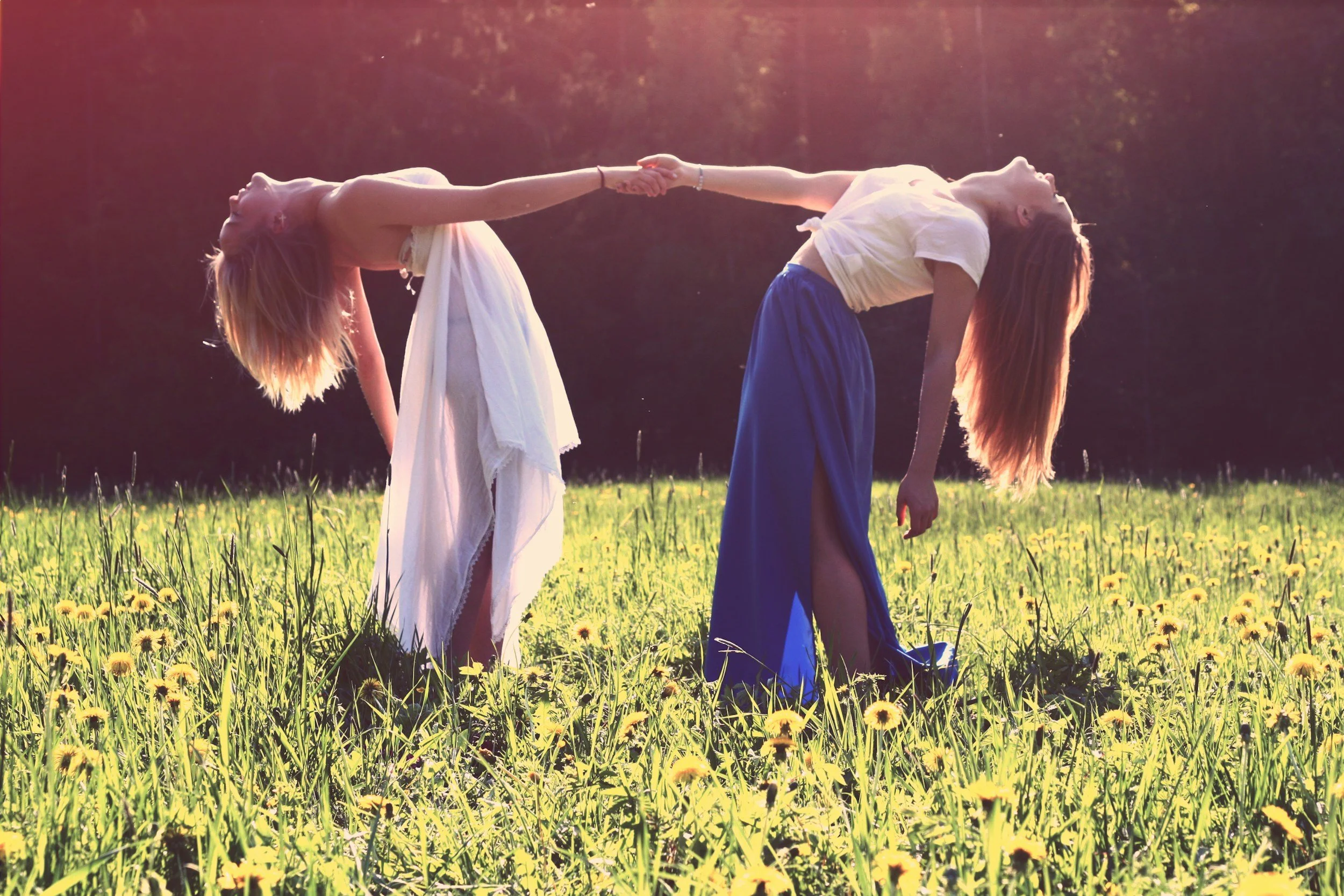Two women in a field holding hands, leaning back, and facing opposite directions during sunset, with a backdrop of trees.
