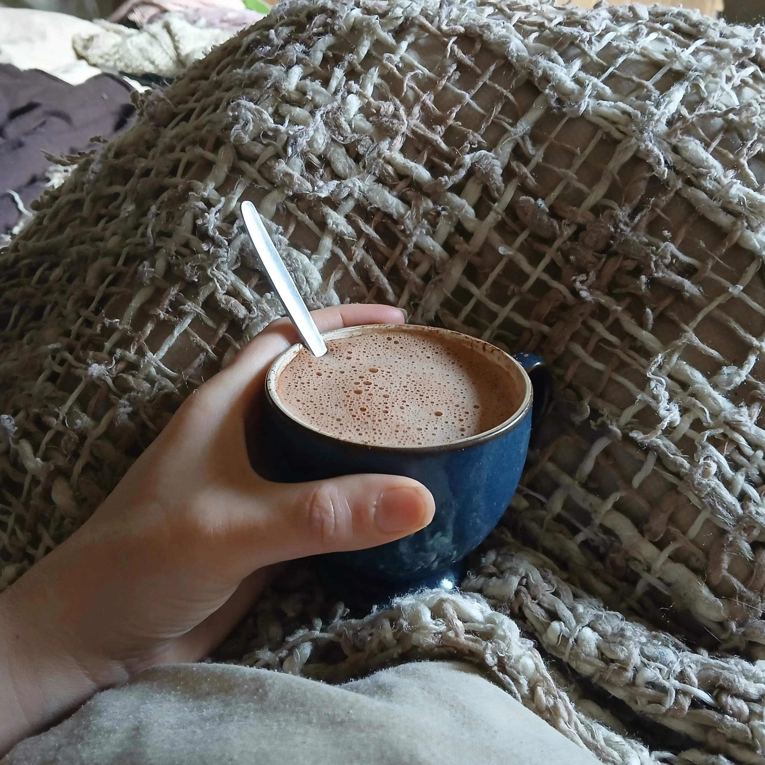 A person's hand holding a blue mug of hot chocolate with foam, resting on a fuzzy, cream-colored blanket.