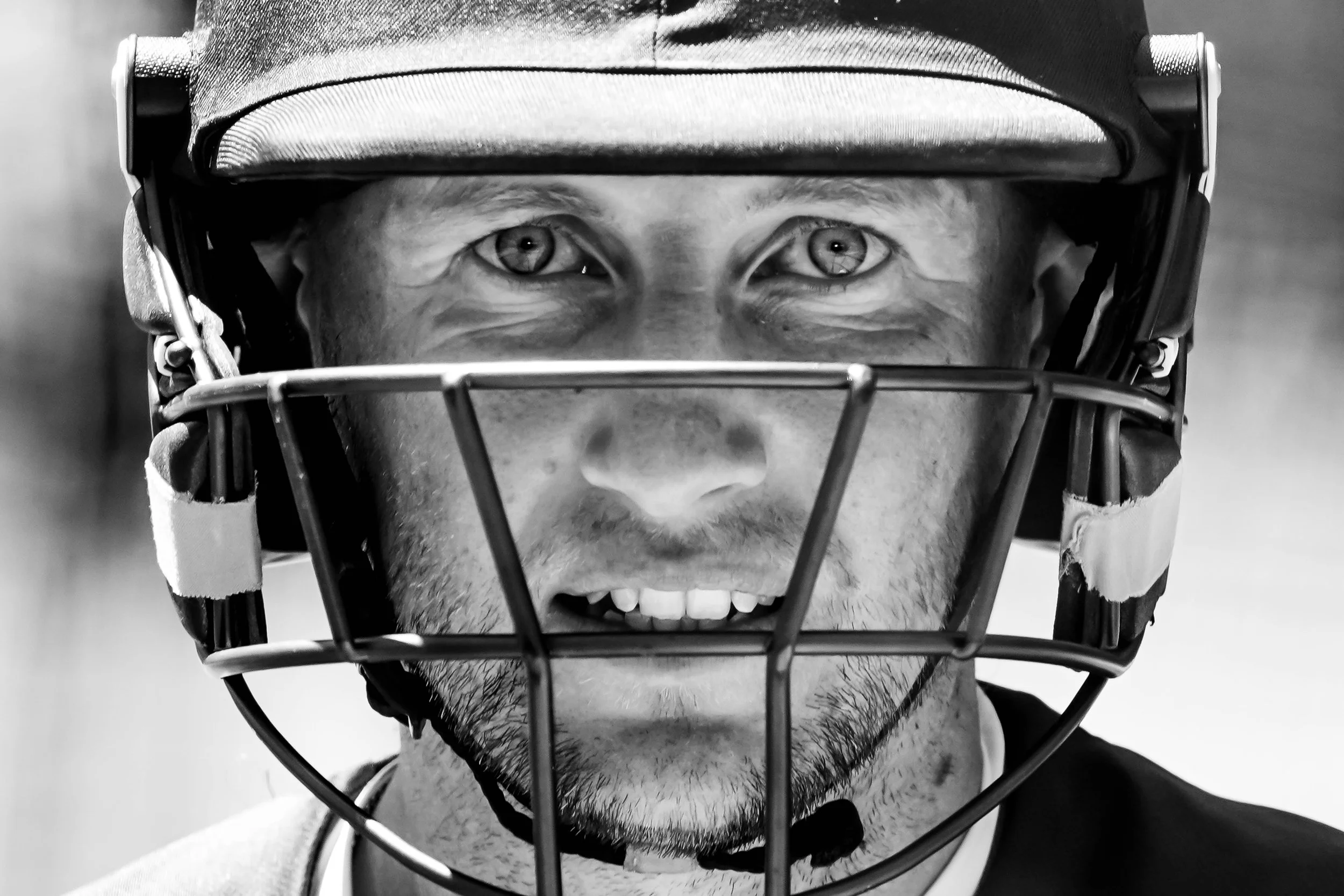 Black and white close-up of a male hockey player wearing a helmet with a face cage, smiling with visible teeth, showing excitement or happiness.