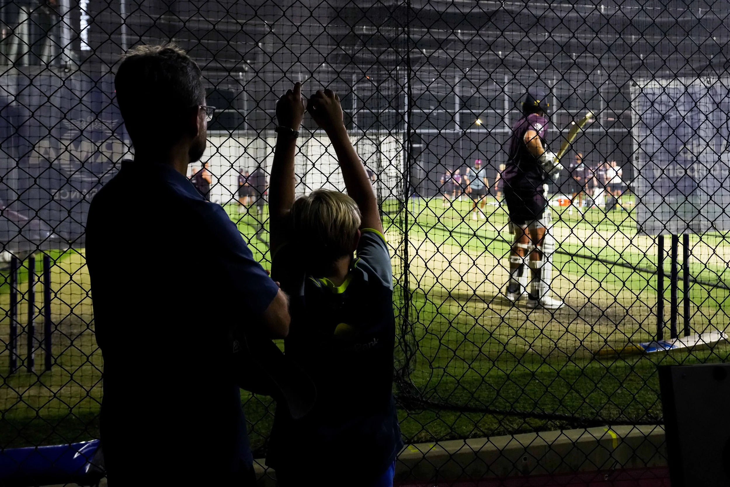 People watching a baseball or softball game through a chain-link fence at night.